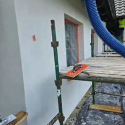 Świeżo otynkowana, biała elewacja domu w Tczewie, widoczna ze stojącego rusztowania z narzędziami murarskimi i niebieskim wężem wentylacyjnym.