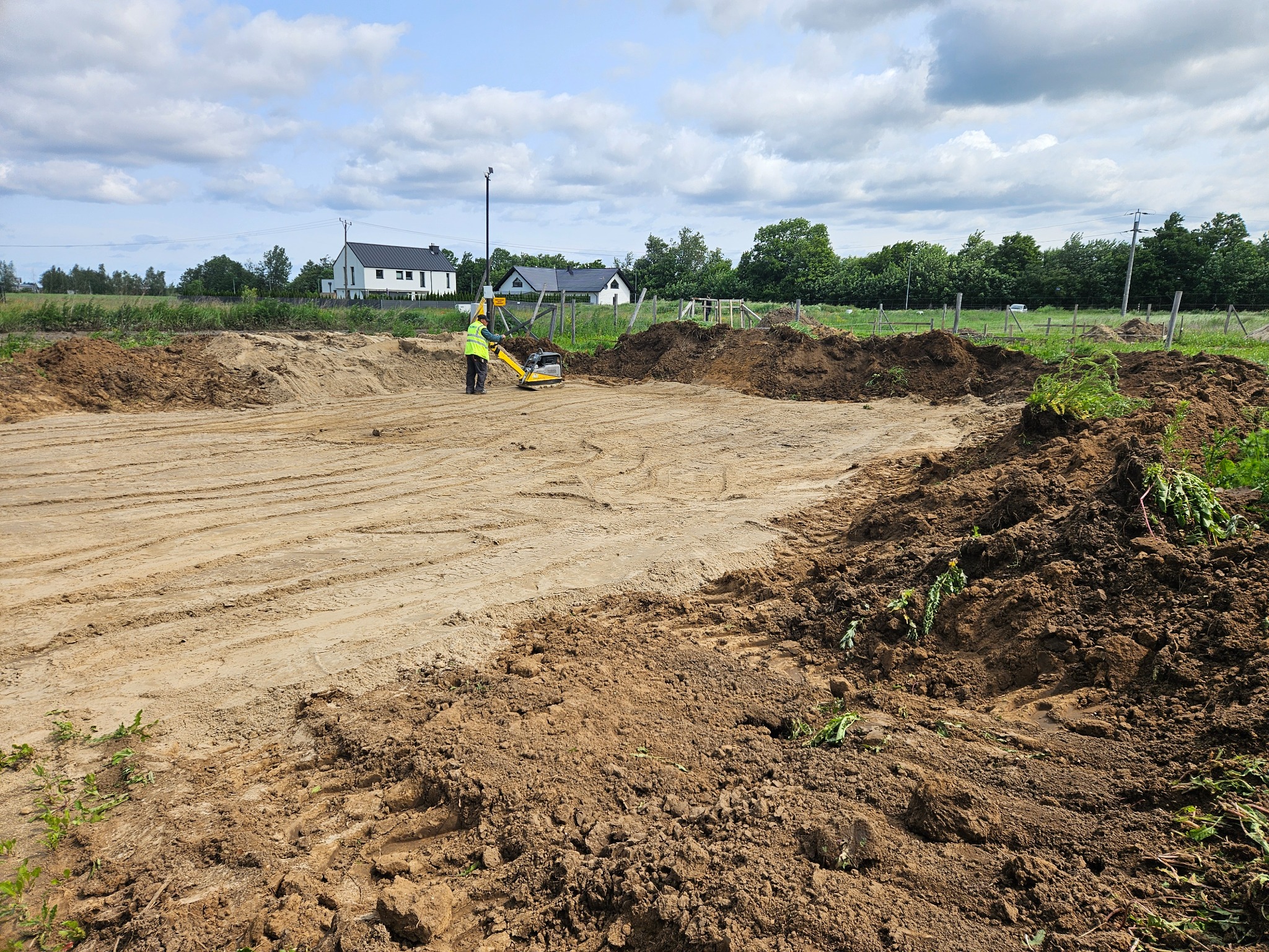 Przygotowanie terenu pod budowę domu: pracownik w żółtej kamizelce zagęszcza piasek zagęszczarką wibracyjną. W tle widoczne domy i błękitne niebo.