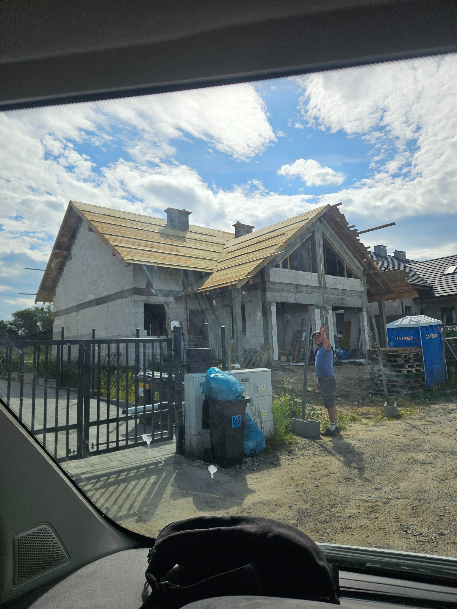 Dom w stanie surowym otwartym z drewnianą konstrukcją dachu, widziany zza szyby samochodu, z pracownikiem budowlanym machającym w tle.