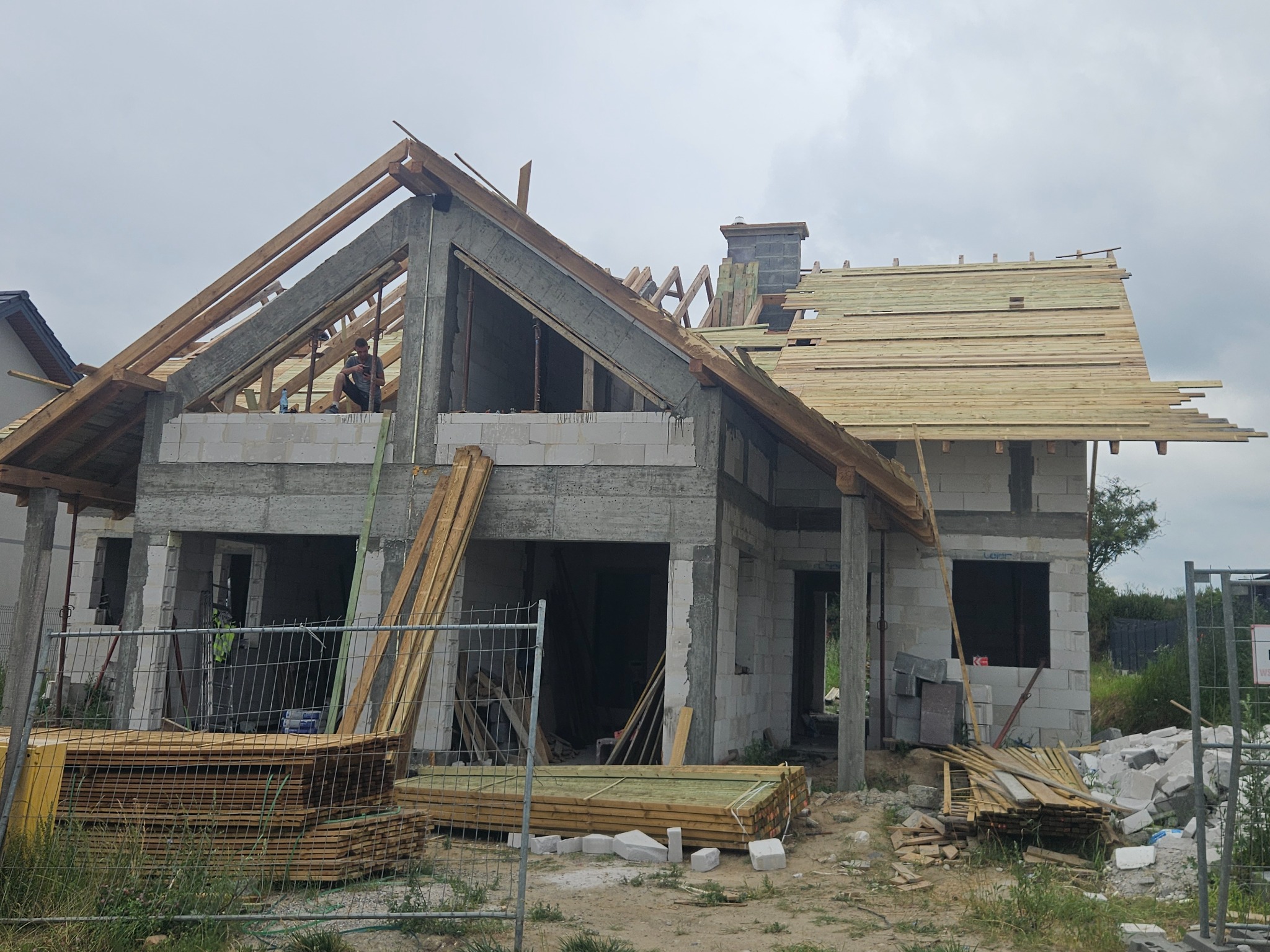 Surowy stan domu z betonu komórkowego w trakcie budowy. Widoczne drewniane krokwie dachu, komin i częściowo ułożone deski na dachu. Na górze budynku siedzi osoba.