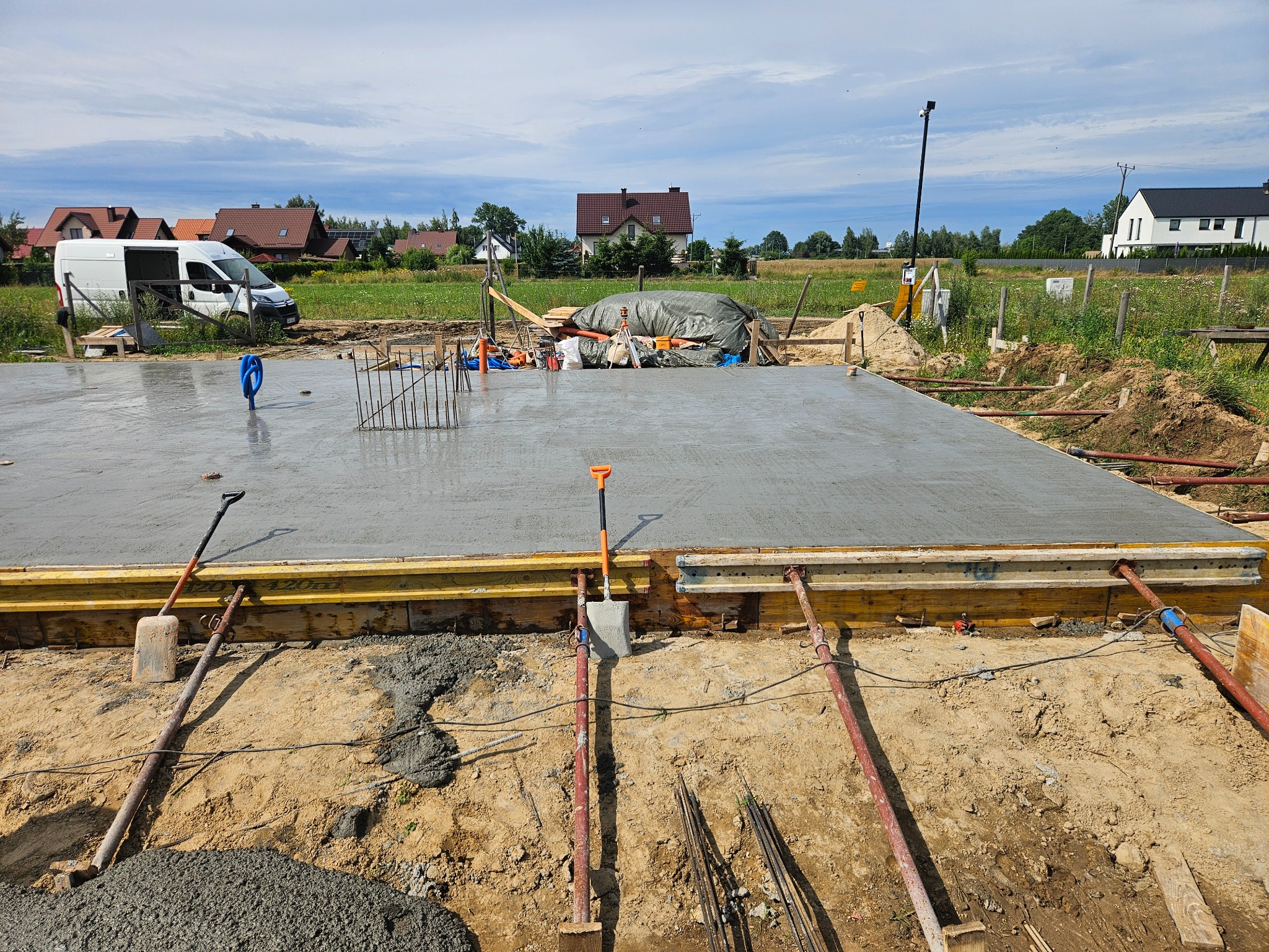 Zalany fundament pod dom jednorodzinny w szalunku, widoczne narzędzia budowlane i zbrojenie. W tle zabudowa jednorodzinna i błękitne niebo.