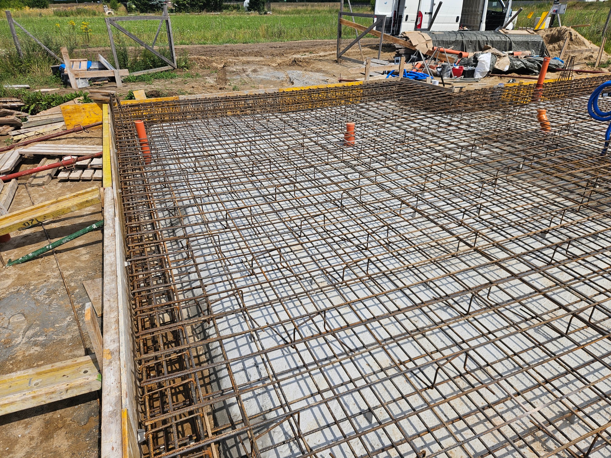 Siatka zbrojeniowa na fundamencie z widocznymi pionowymi rurami i żółtym szalunkiem, przygotowana do zalania betonem. Zbliżenie na konstrukcję.