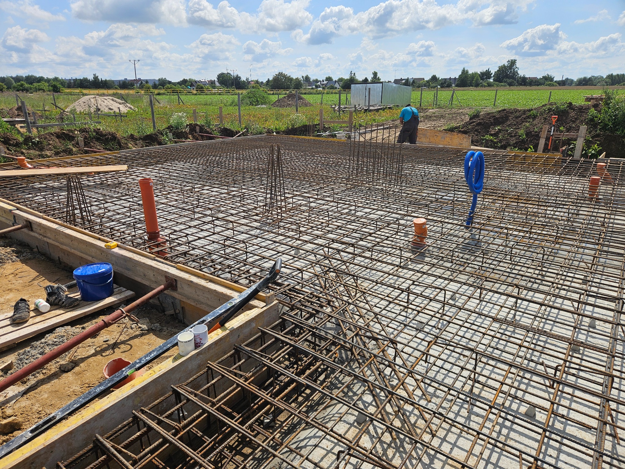 Zbrojenie fundamentów domu jednorodzinnego z widocznymi elementami szalunku i instalacji. W tle krajobraz wiejski i pracownik budowlany.