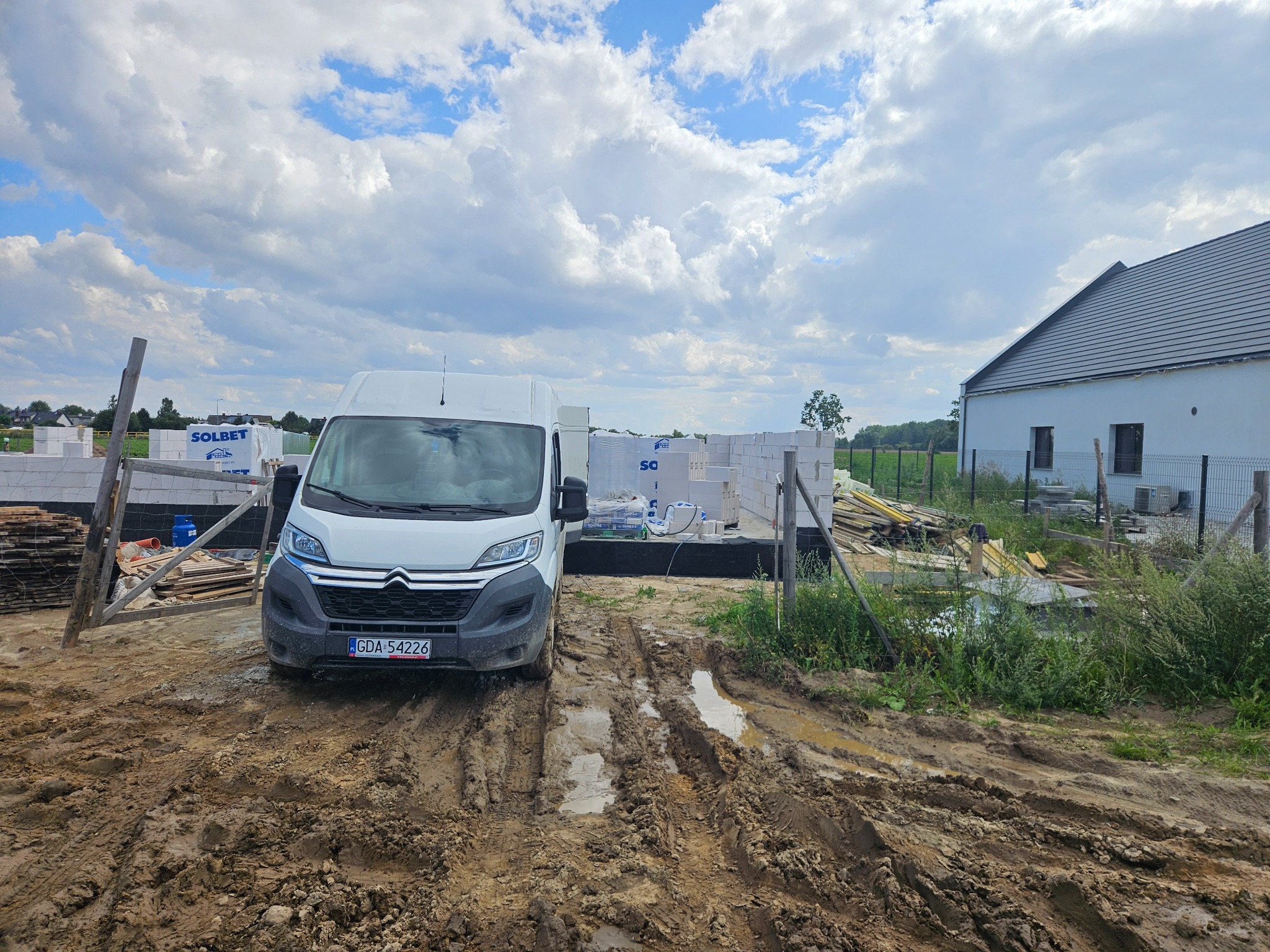 Budowa domu w trakcie murowania ścian, widoczny biały bus dostawczy na błotnistym terenie. Ściany wzniesione z białych bloczków, palety z materiałami budowlanymi w tle.