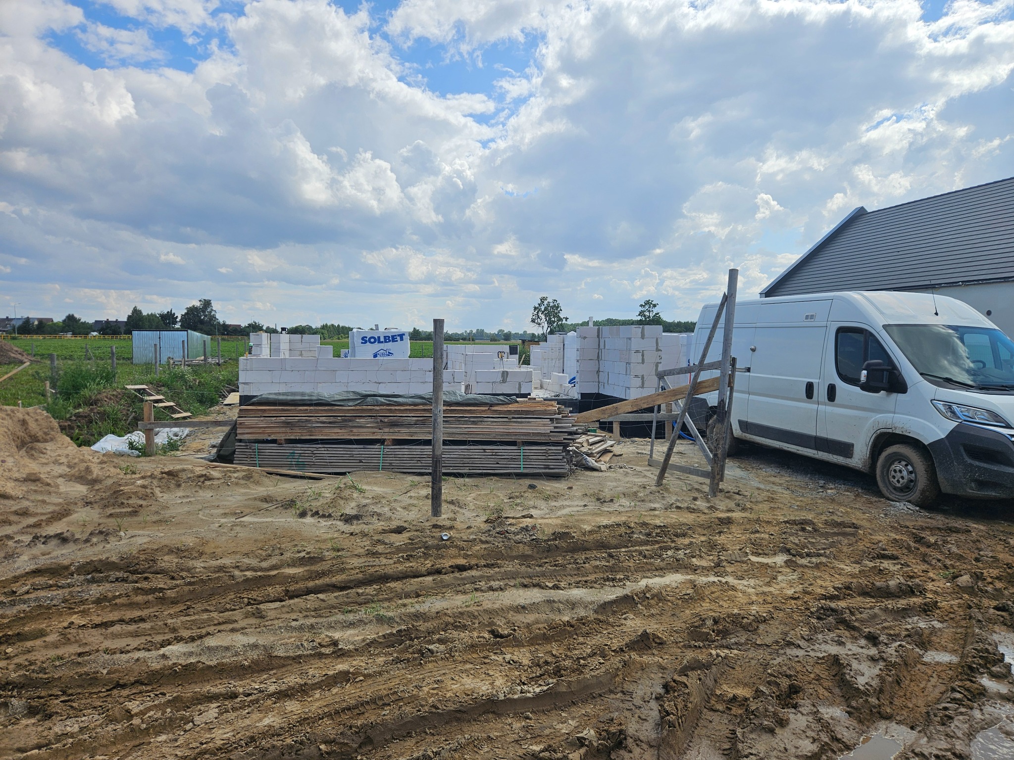 Budowa domu w trakcie: białe bloczki Solbet, drewniane deski, teren budowy z błotem i biały bus w tle, pochmurne niebo. Widoczny fragment dachu sąsiedniego budynku.