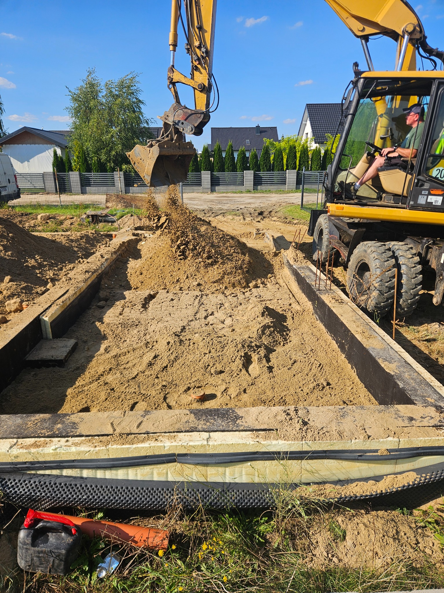 Żółta koparka zasypuje piaskiem przygotowany fundament z izolacją. Widoczne zbrojenia i elementy szalunku. W tle zabudowa jednorodzinna i błękitne niebo.