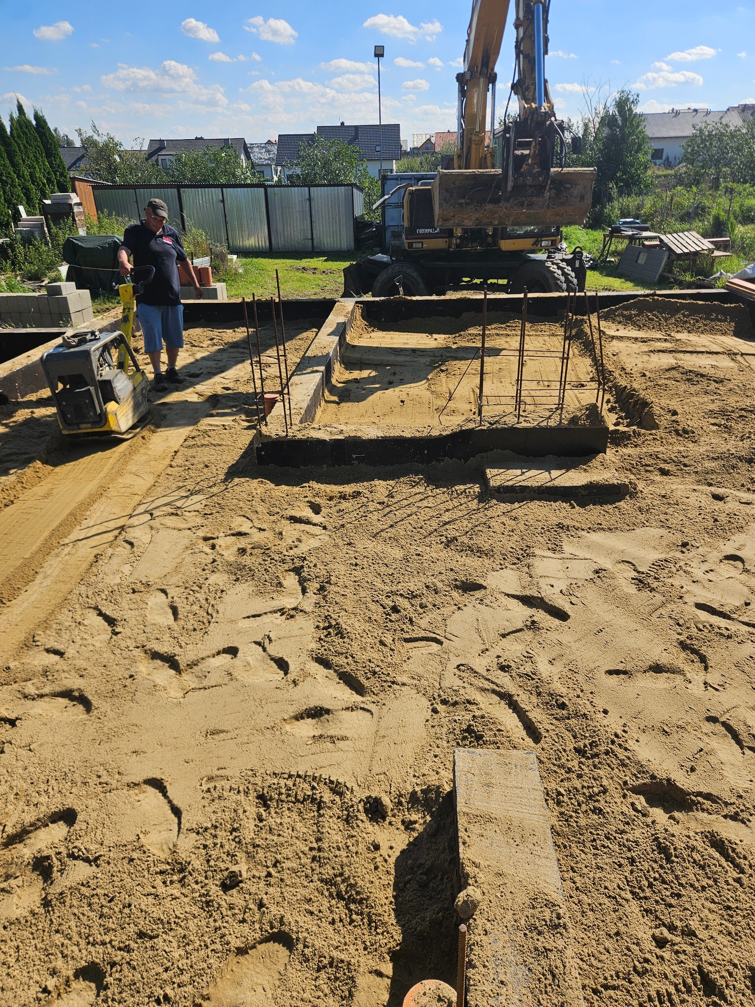 Prace ziemne: zagęszczarka w akcji, przygotowanie fundamentów pod budowę. Widoczny wykop, zbrojenie i koparka w tle, sugerujące kompleksową realizację.