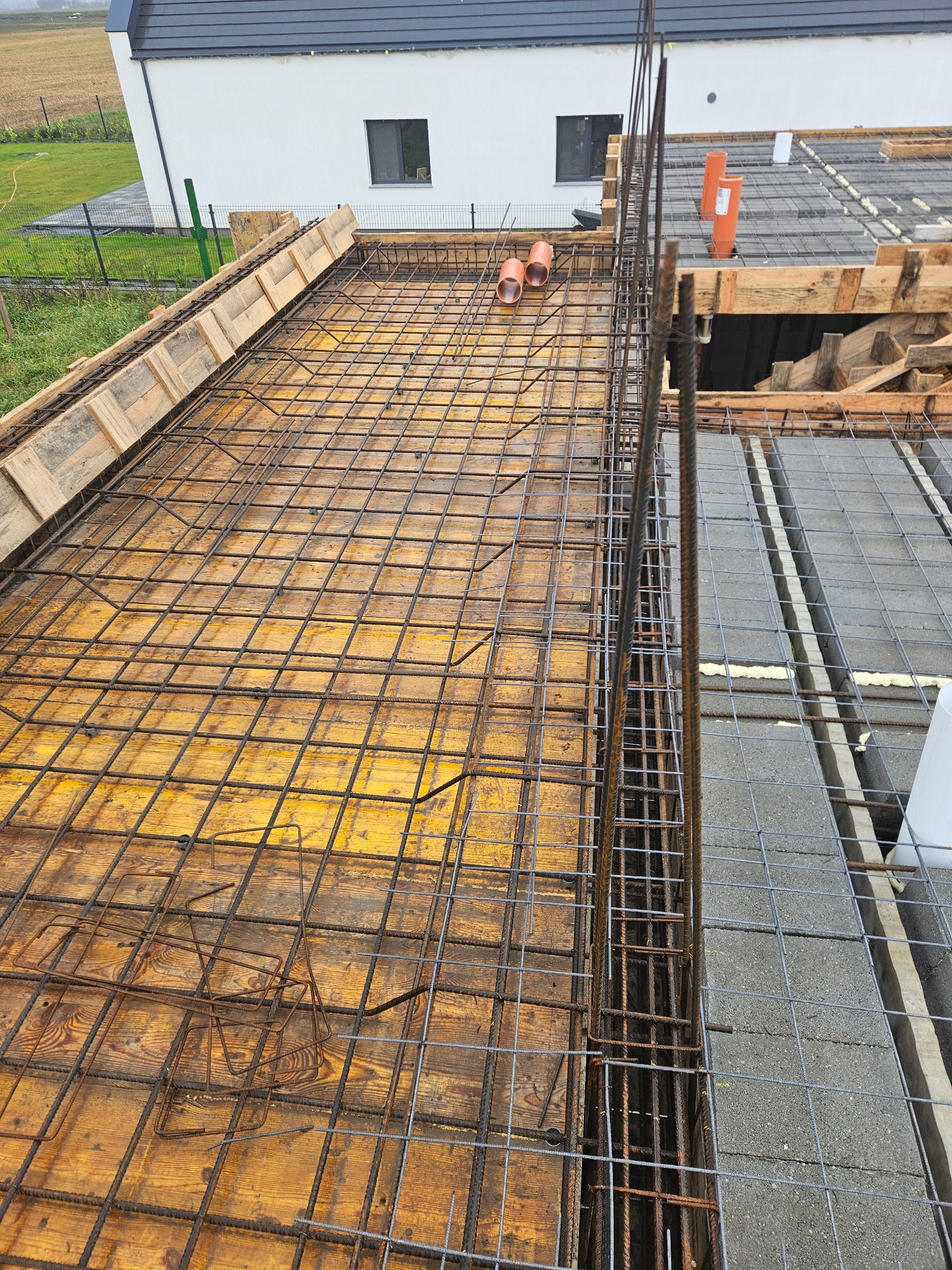 Zbrojenie stropu na deskowaniu z widocznymi elementami instalacji. Stalowe pręty tworzą siatkę, przygotowane do zalania betonem. W tle budynek w trakcie budowy.