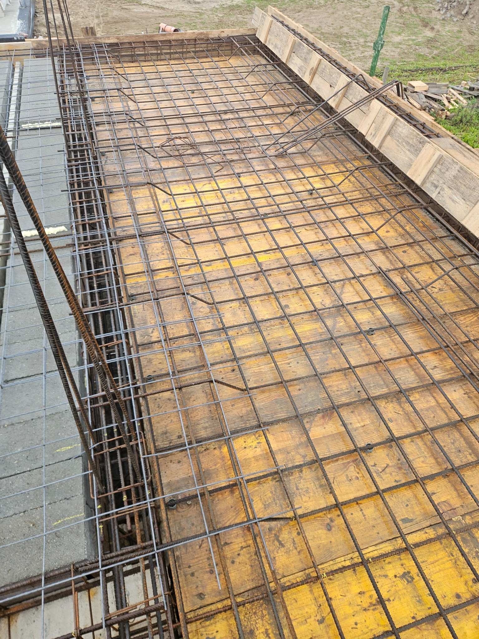 Zbrojenie stropu na deskowaniu z żółtej sklejki. Widoczne pręty zbrojeniowe tworzące siatkę, zbrojenie pionowe. Grunt w tle.