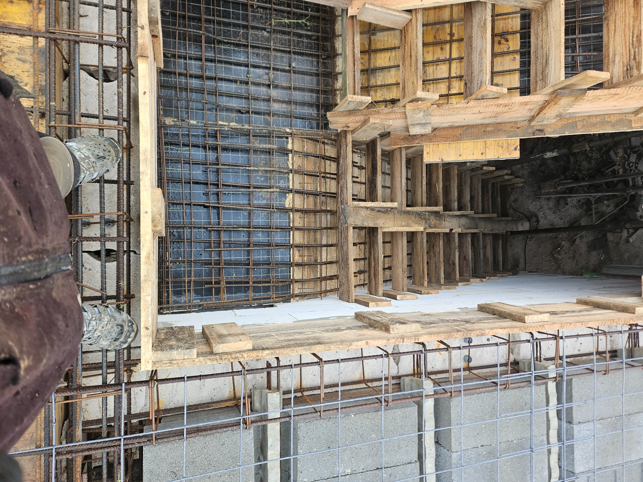 Zbrojenie i szalunek fundamentów domu z widoczną siatką zbrojeniową, drewnianą konstrukcją szalunku oraz fragmentem buta osoby nadzorującej prace.