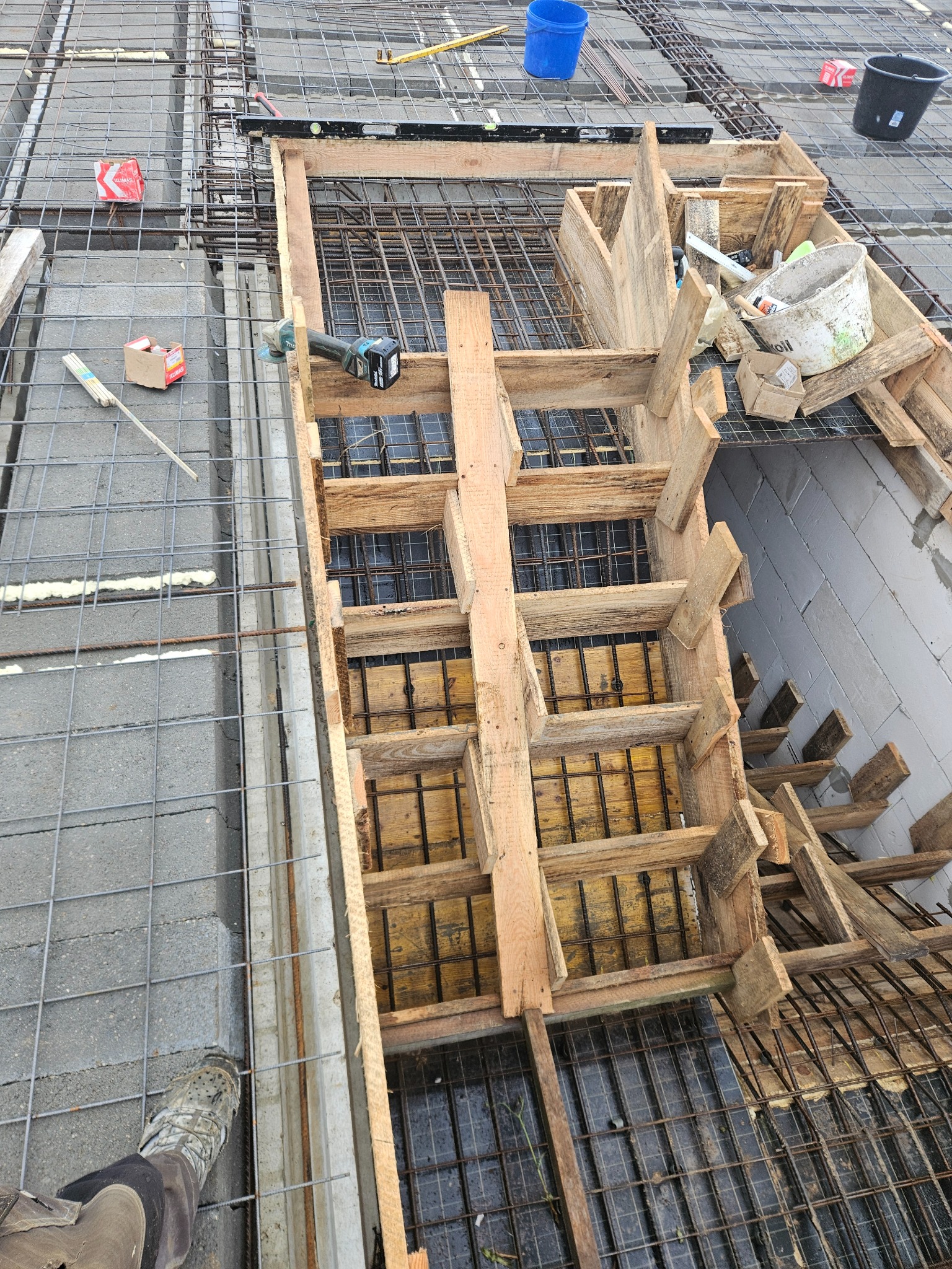 Zbrojenie stropu z drewnianym szalunkiem, widoczne pręty zbrojeniowe i elementy konstrukcji. Na pierwszym planie fragment buta pracownika.