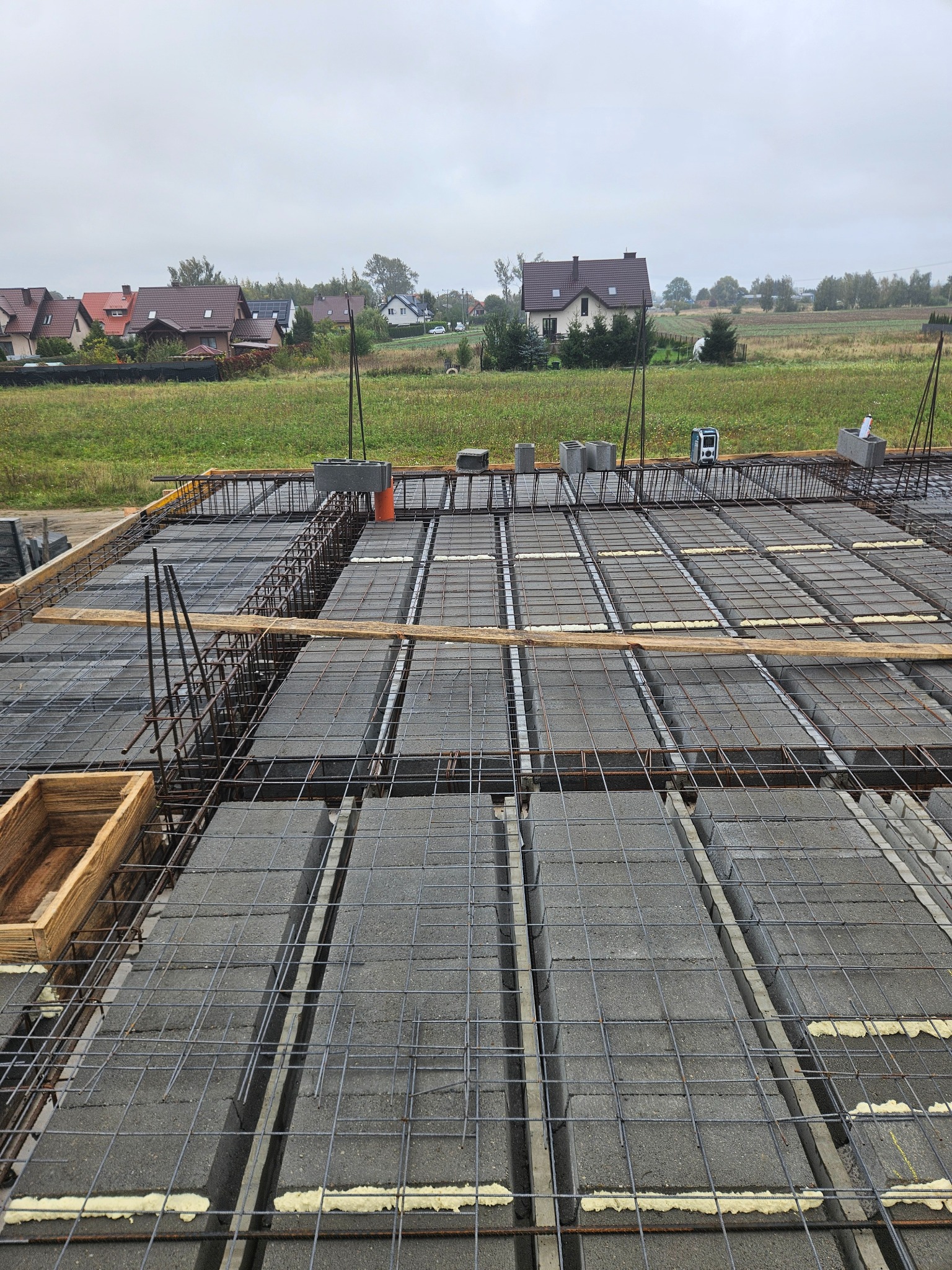 Zbrojenie stropu Teriva z pustakami szalunkowymi, widoczne pręty zbrojeniowe i pianka montażowa. W tle domy jednorodzinne i pochmurne niebo.