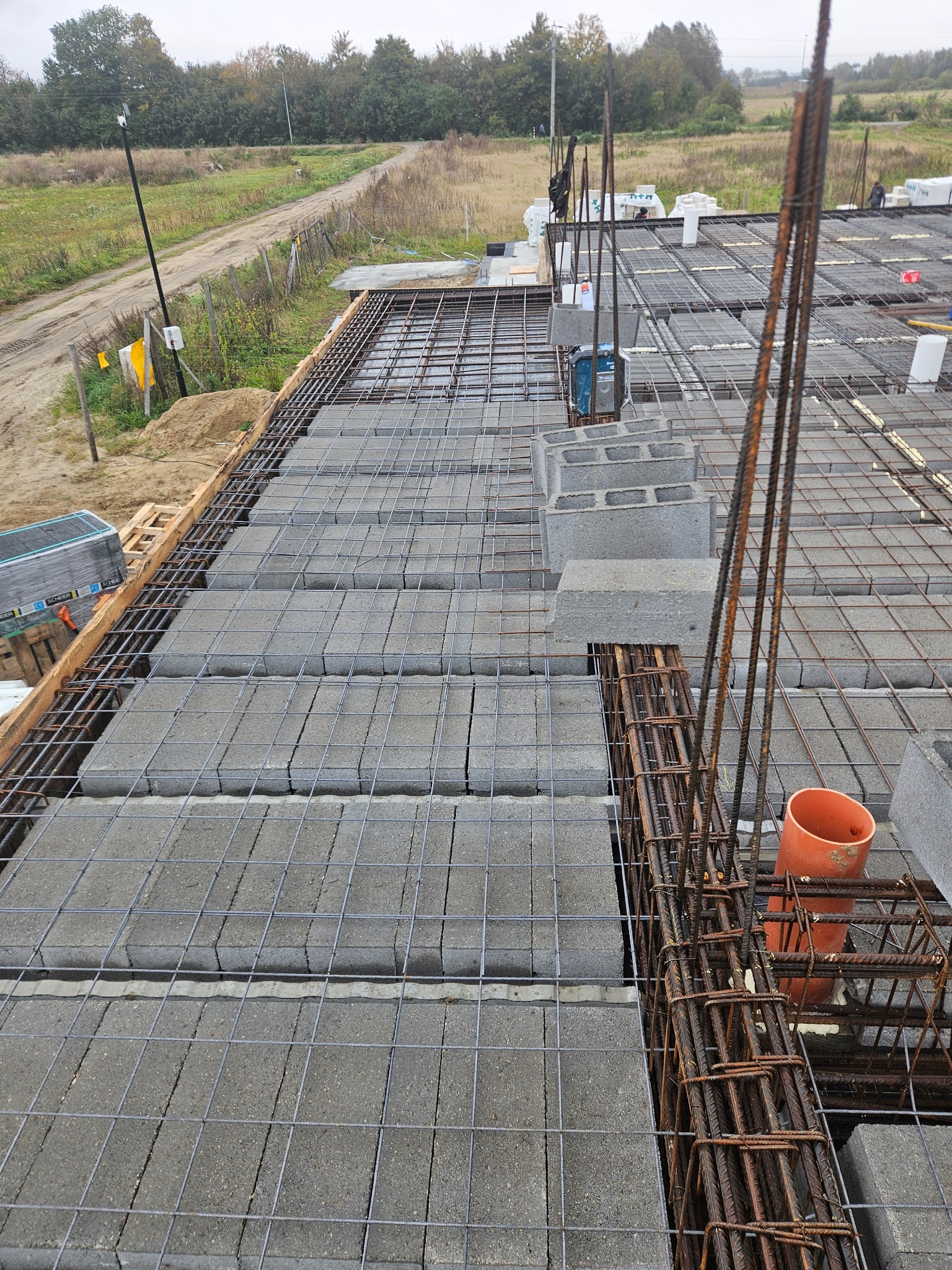 Zbrojenie stropu Teriva z pustaków szalunkowych i wieńcem. Widoczne pręty zbrojeniowe pionowe i poziome oraz pomarańczowa rura kanalizacyjna. Budowa domu w trakcie realizacji.