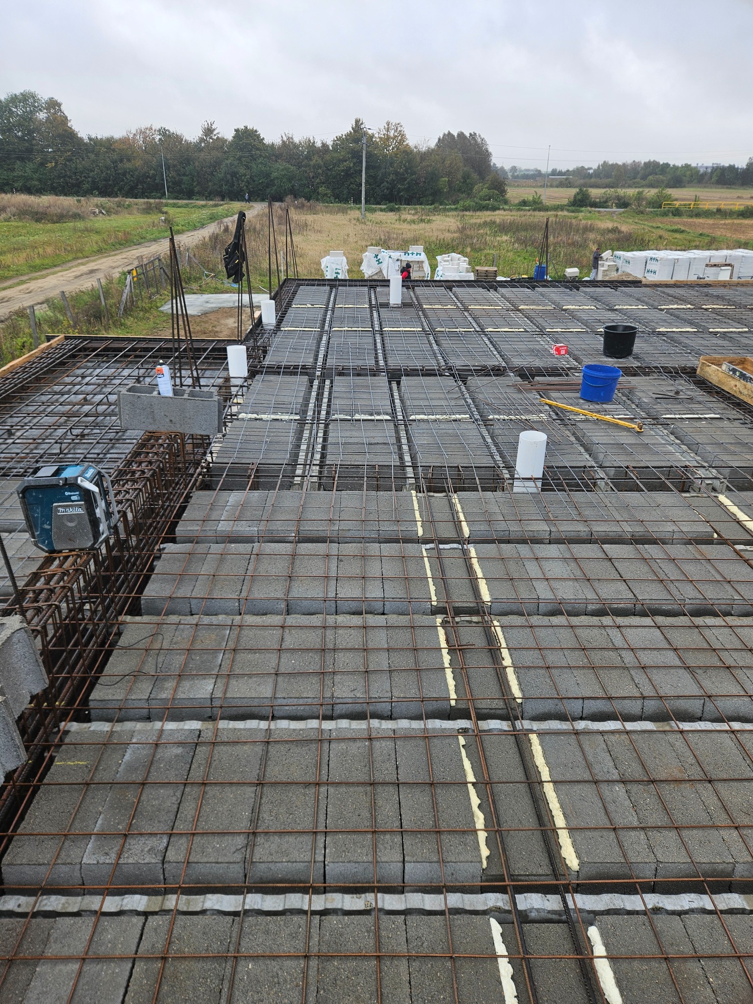 Budowa stropu: szalunek zbrojony siatką i bloczkami betonowymi, widoczne narzędzia i materiały budowlane na tle krajobrazu. Etap prac w toku.