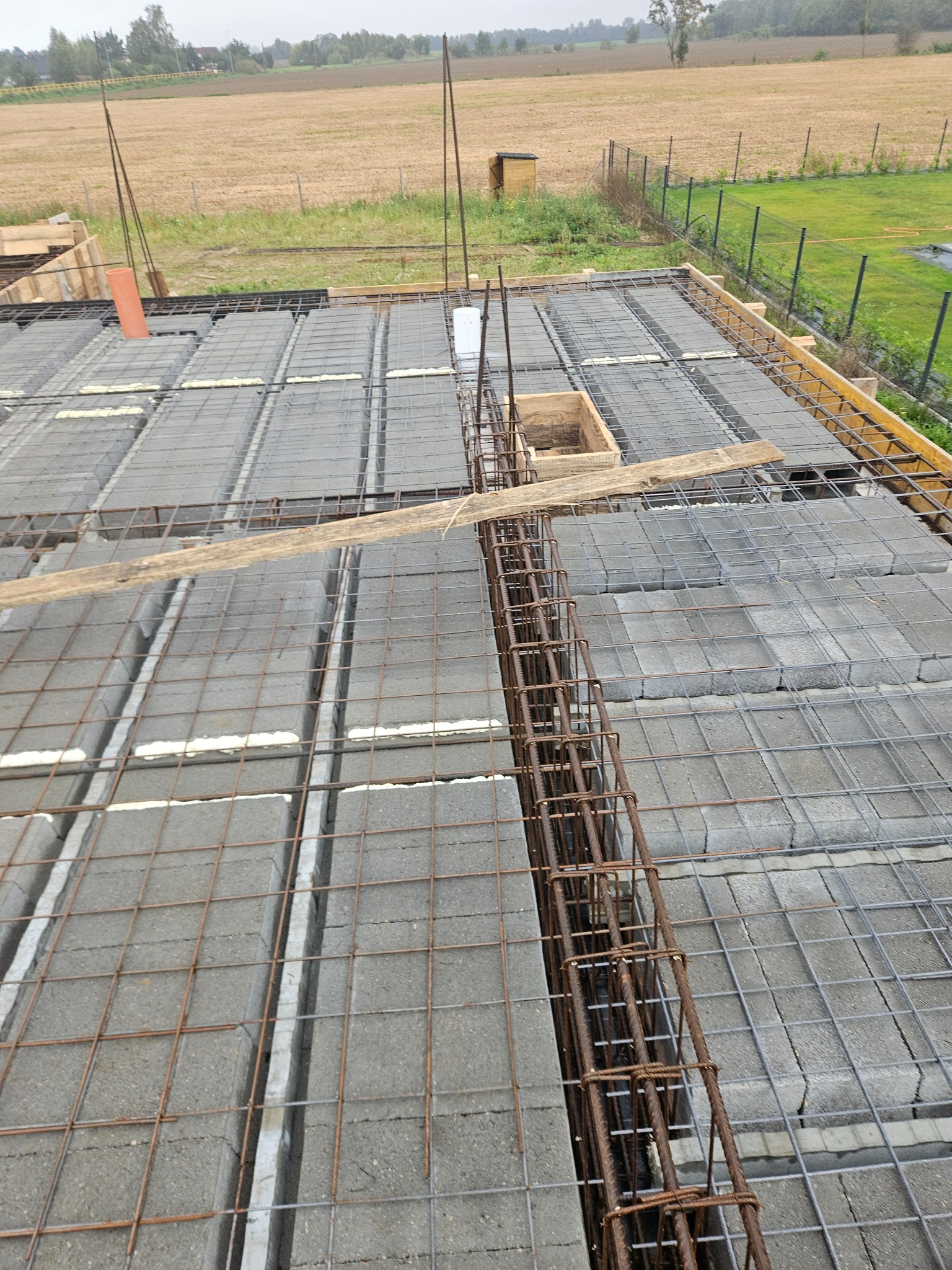 Zbrojenie stropu Teriva z pustaków szalunkowych, widok z góry na siatkę zbrojeniową i wystające pręty. W tle pole i zabudowania.