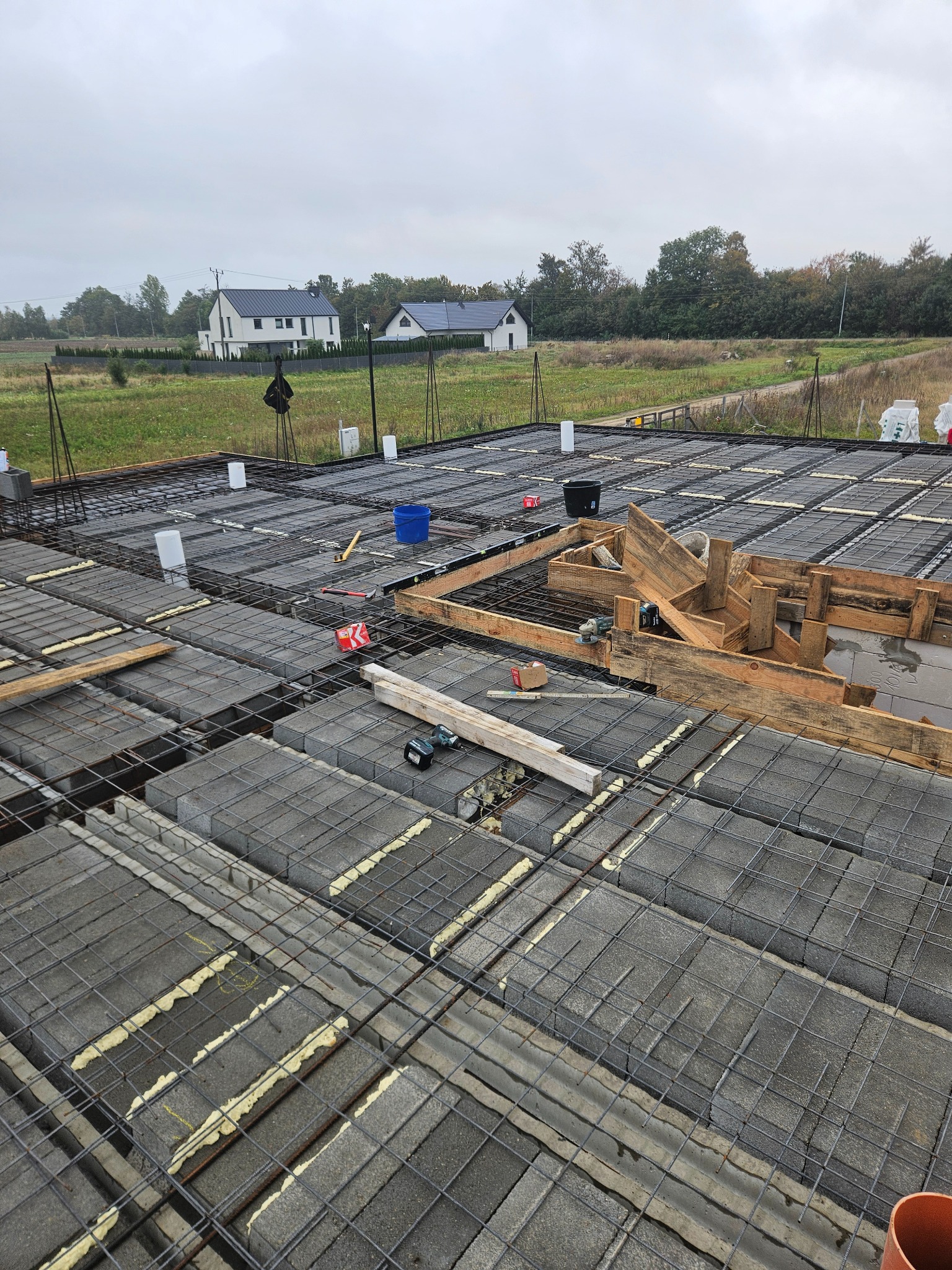 Budowa stropu Teriva: szare bloczki, zbrojenie stalowe, pianka montażowa. Widoczne narzędzia, szalunki i domy w tle. Ujęcie z góry na plac budowy w pochmurny dzień.