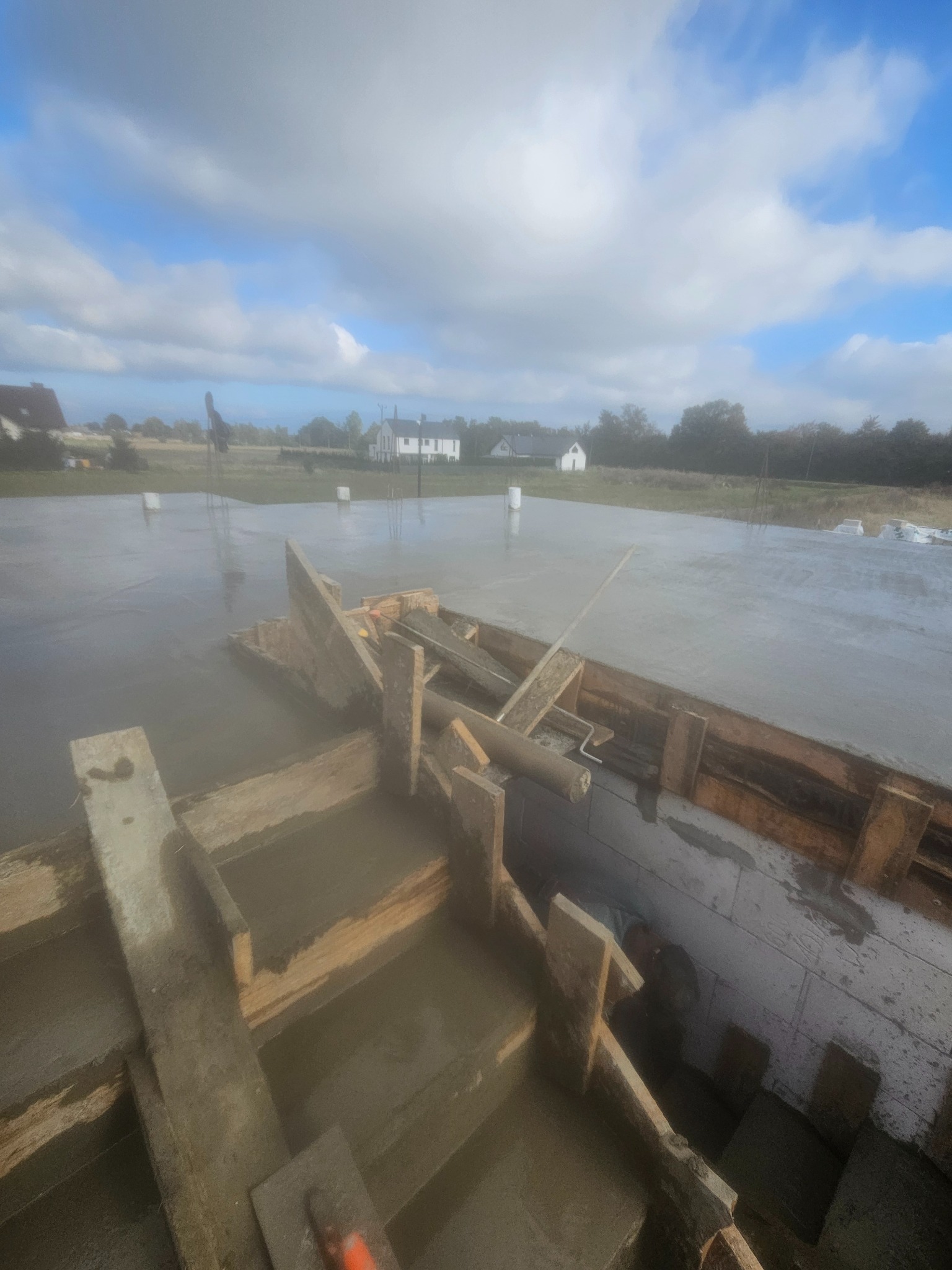 Wylana, świeża betonowa posadzka w szalunku z drewnianych desek na budowie domu jednorodzinnego. W tle błękitne niebo i zabudowania.