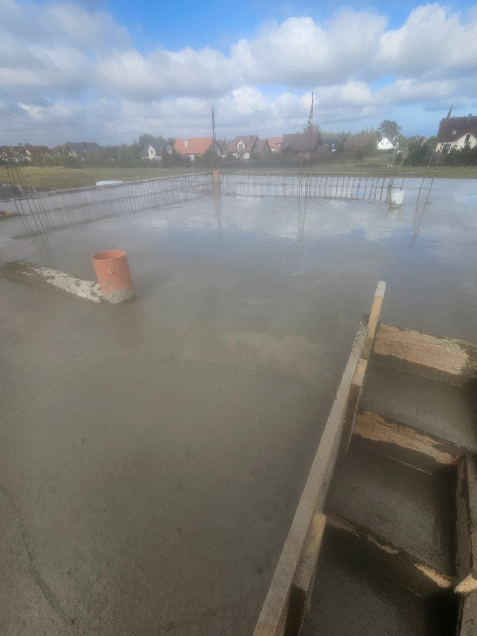 Świeżo wylana płyta fundamentowa z widocznym zbrojeniem i rurą kanalizacyjną na tle zabudowy jednorodzinnej pod błękitnym niebem.