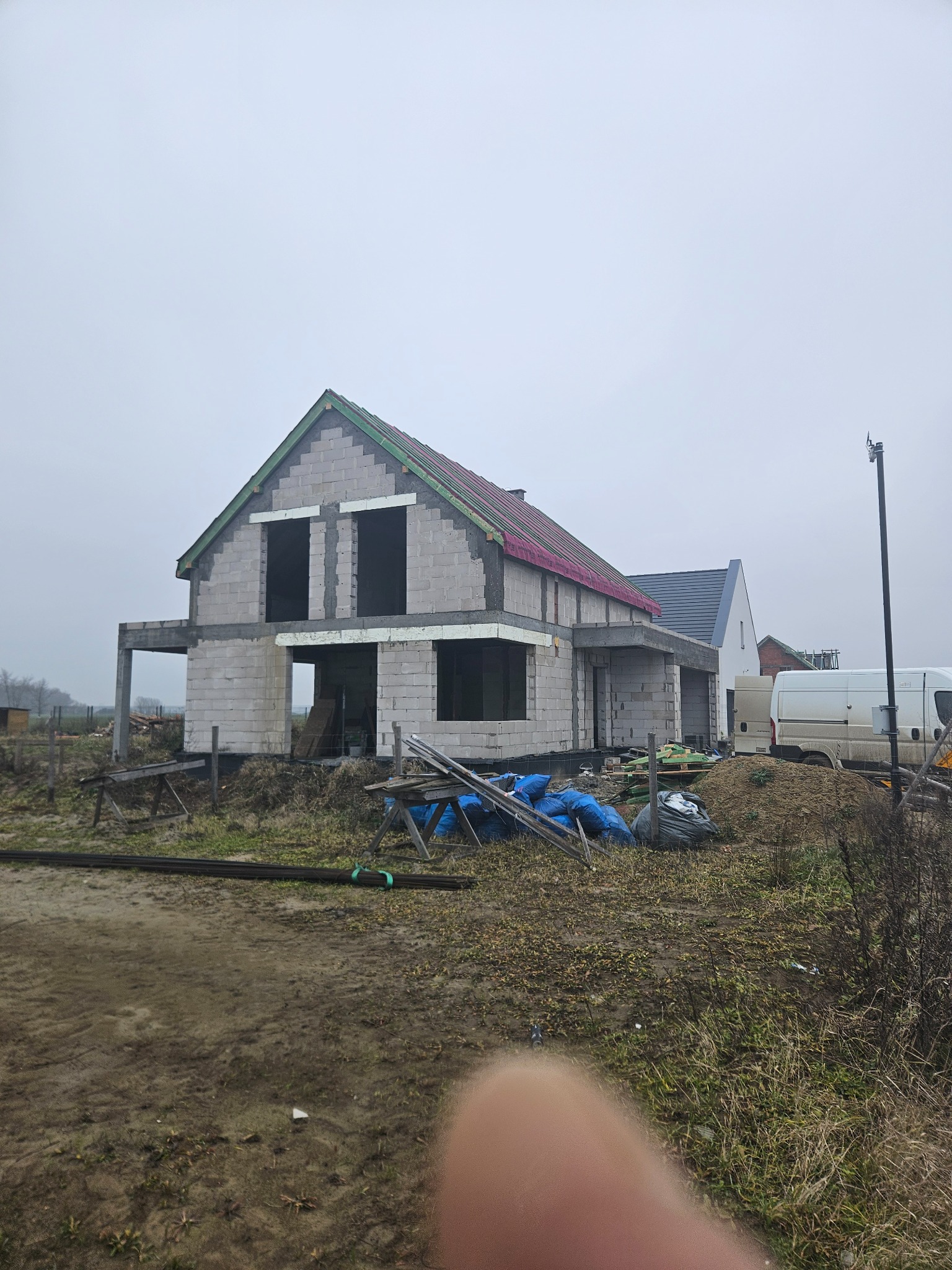 Niedokończony dom z betonu komórkowego w trakcie budowy, widoczne zbrojenia dachu i okna bez ram. Na pierwszym planie teren budowy i materiały budowlane.