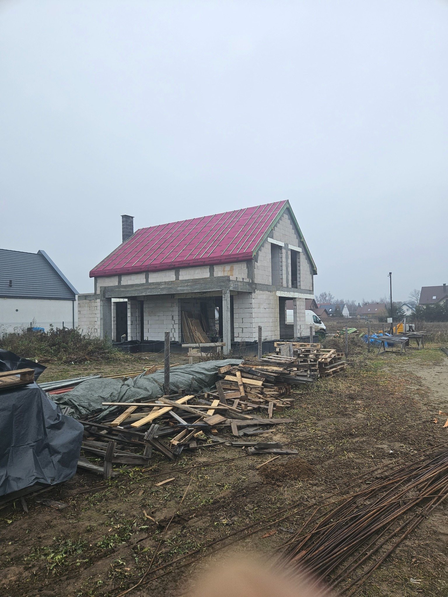 Surowy dom z czerwonym dachem w trakcie budowy, widoczne białe bloczki, drewniane elementy i zbrojenia na błotnistym terenie. Pochmurne niebo w tle.