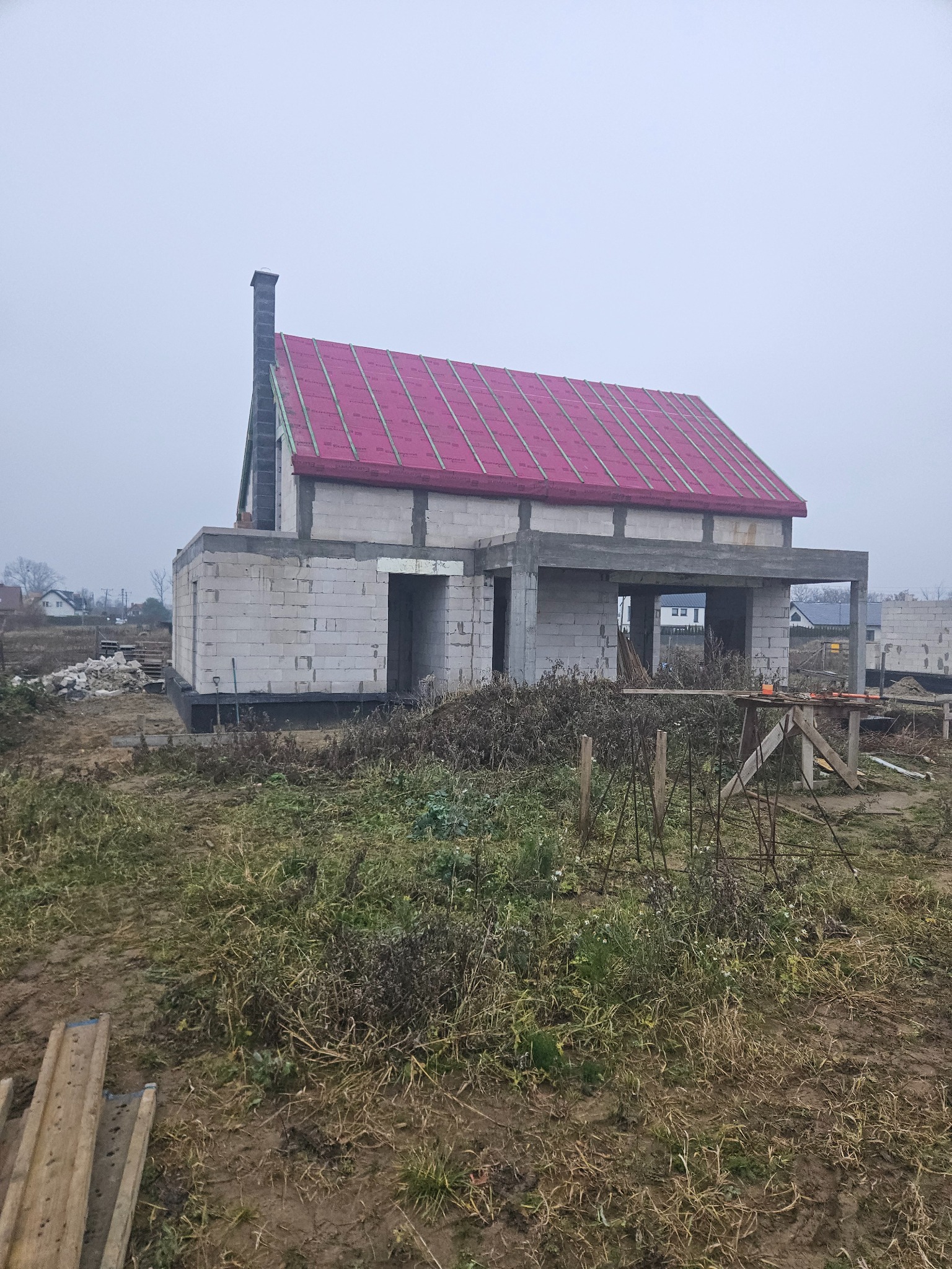 Surowy stan domu z białego pustaka z czerwonym dachem, kominem i otworami okiennymi, otoczony niezagospodarowanym terenem w pochmurny dzień.