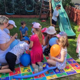 ANIMKA Człuchów - Animator z grupą dzieci siedzących na kolorowej macie edukacyjnej w ogrodzie, przygotowuje balony na przyjęcie urodzinowe. W tle zjeżdżalnia i huśtawki.