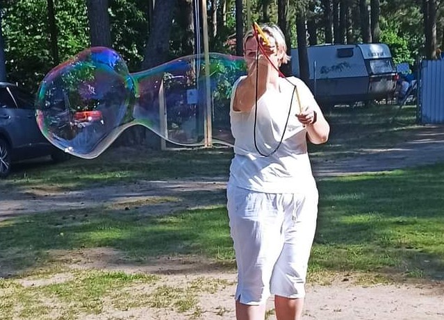 Kobieta w białym ubraniu puszcza ogromne bańki mydlane na trawniku w Człuchowie, w tle drzewa i przyczepa kempingowa.