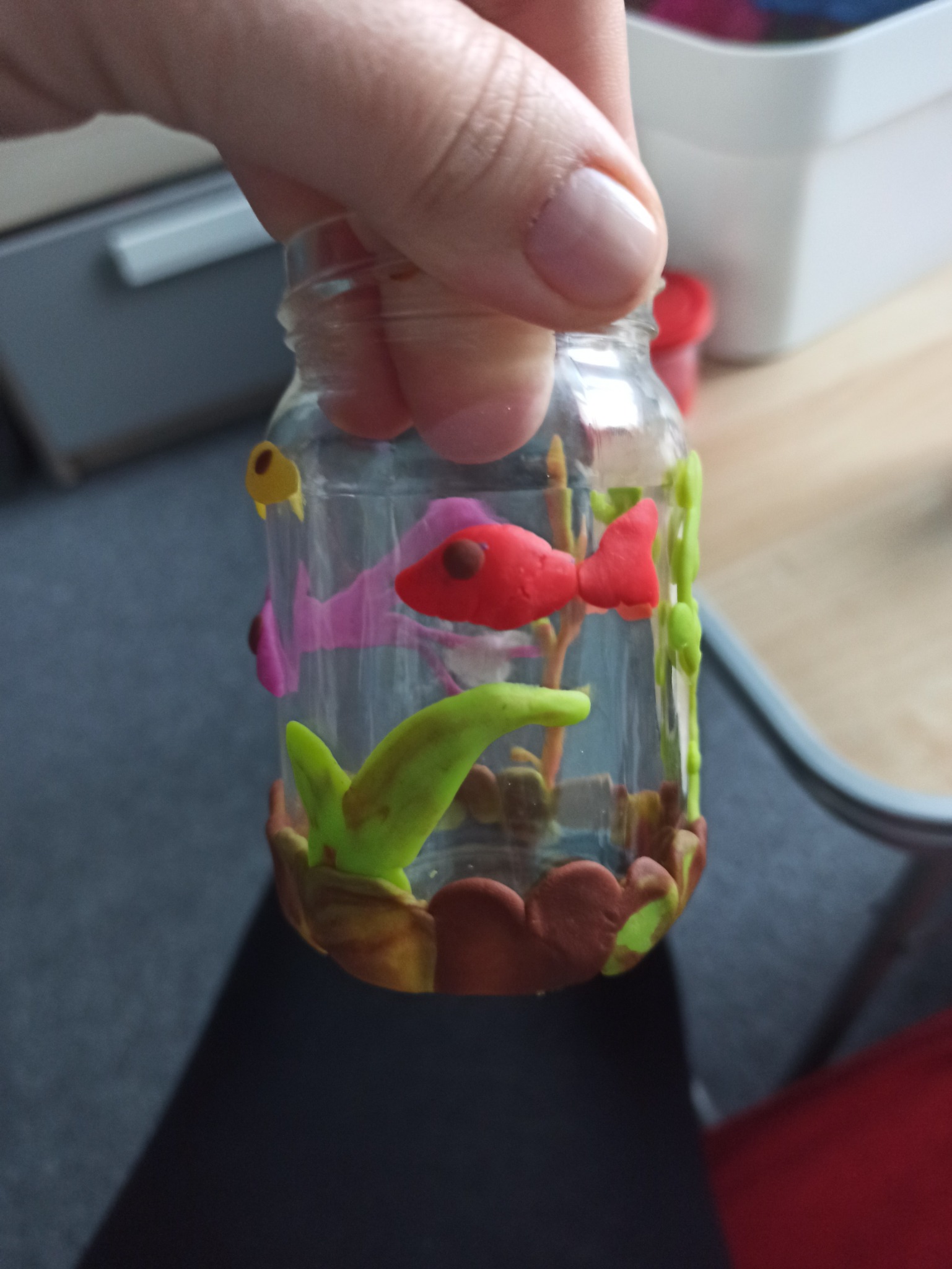 Ręcznie zdobiony słoik z motywem akwarium z plasteliny, przedstawiający kolorowe rybki i rośliny, trzymany w dłoni.