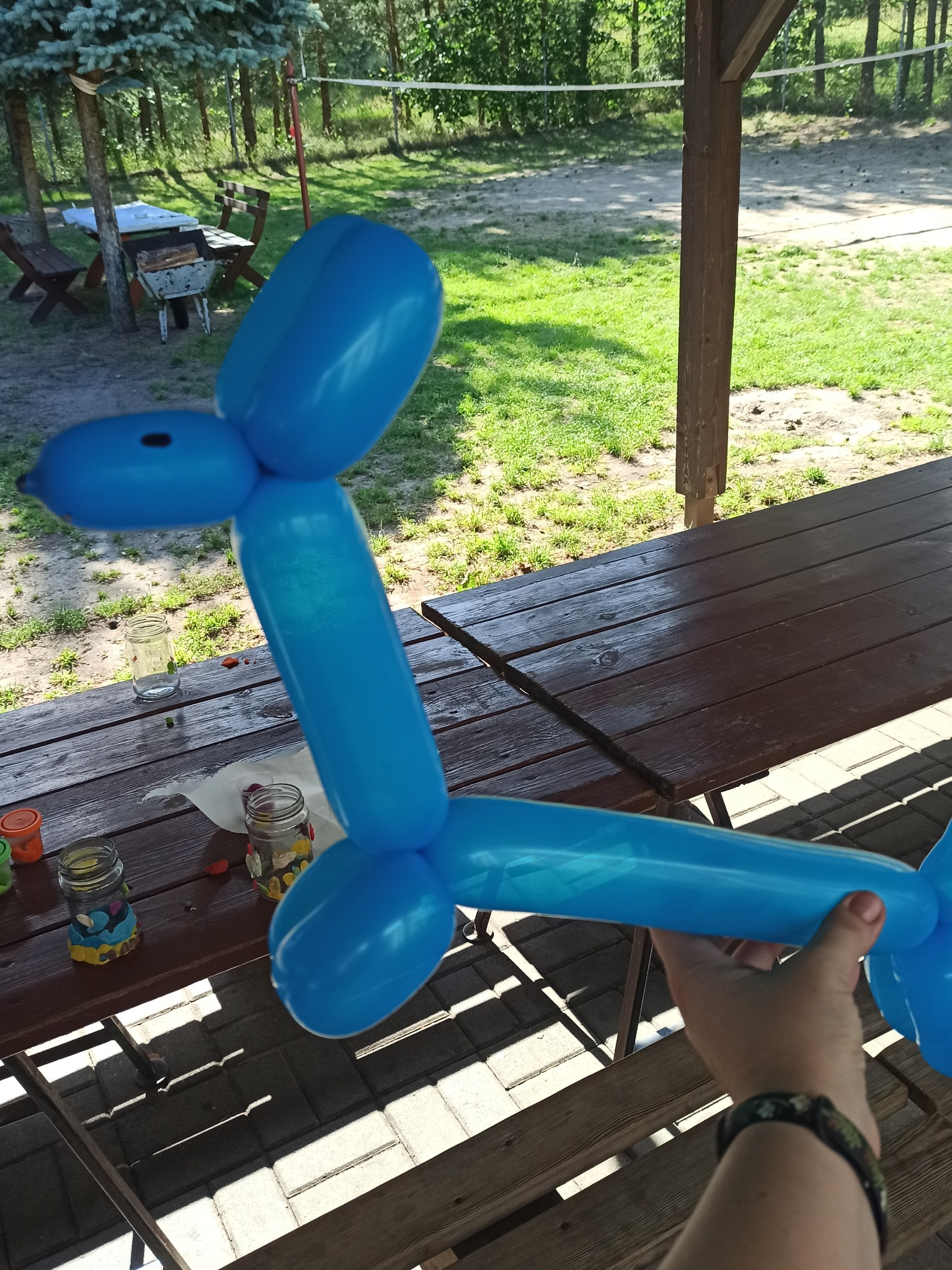 Niebieski balon w kształcie zwierzęcia trzymany w ręku na tle drewnianego stołu z dekoracjami w słoikach, plenerowa sceneria.