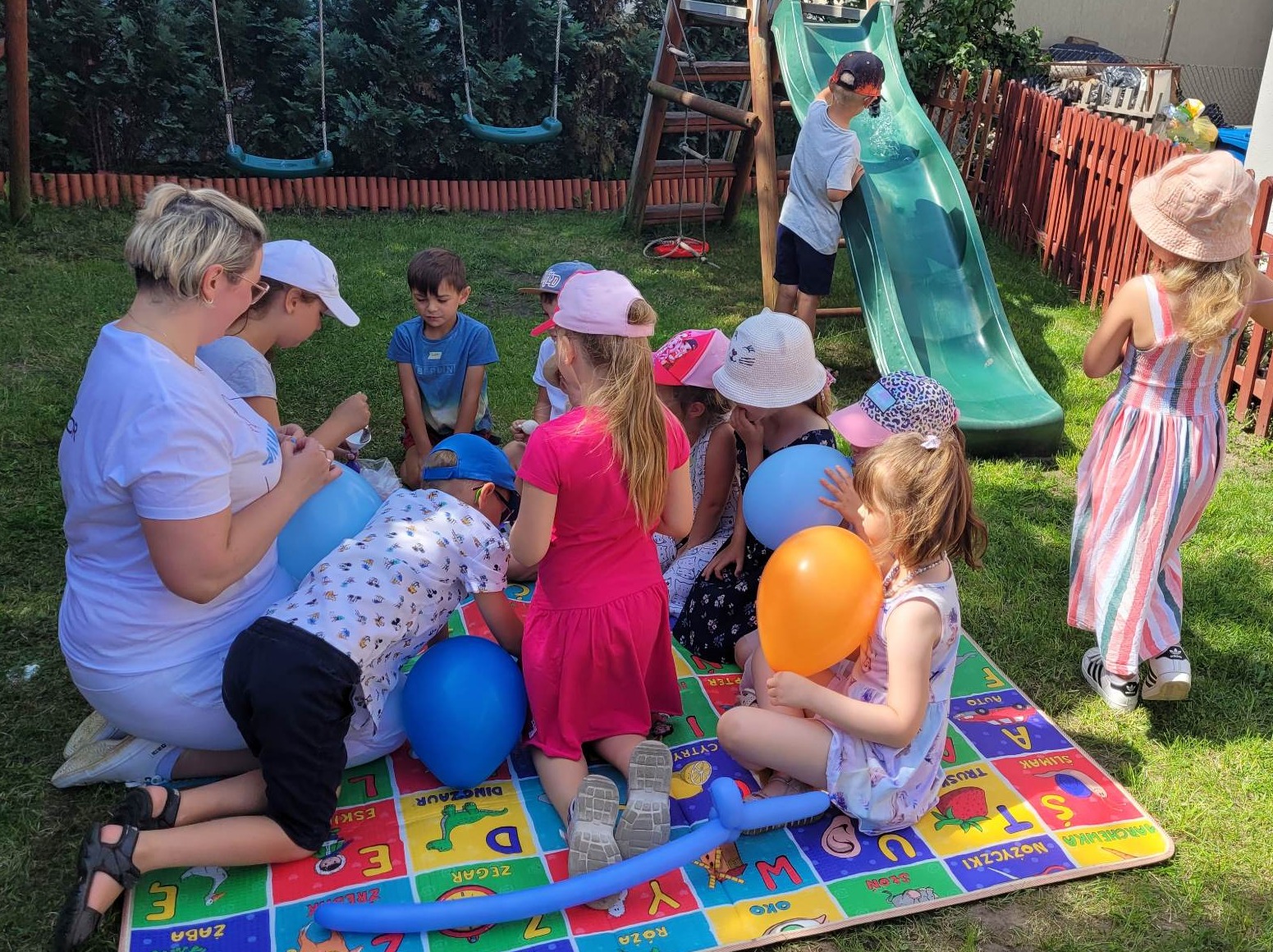 Animator z grupą dzieci siedzących na kolorowej macie edukacyjnej w ogrodzie, przygotowuje balony na przyjęcie urodzinowe. W tle zjeżdżalnia i huśtawki.