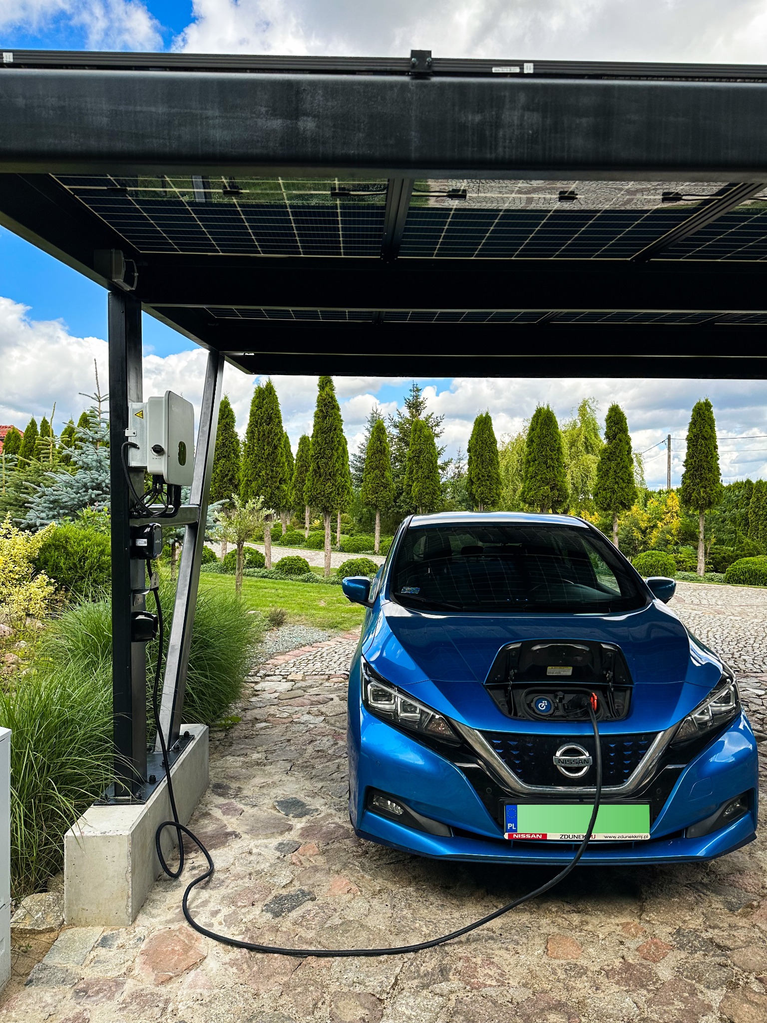 Niebieski samochód elektryczny ładowany pod wiatą fotowoltaiczną w ogrodzie, widoczny falownik na słupie wiaty i brukowana nawierzchnia.