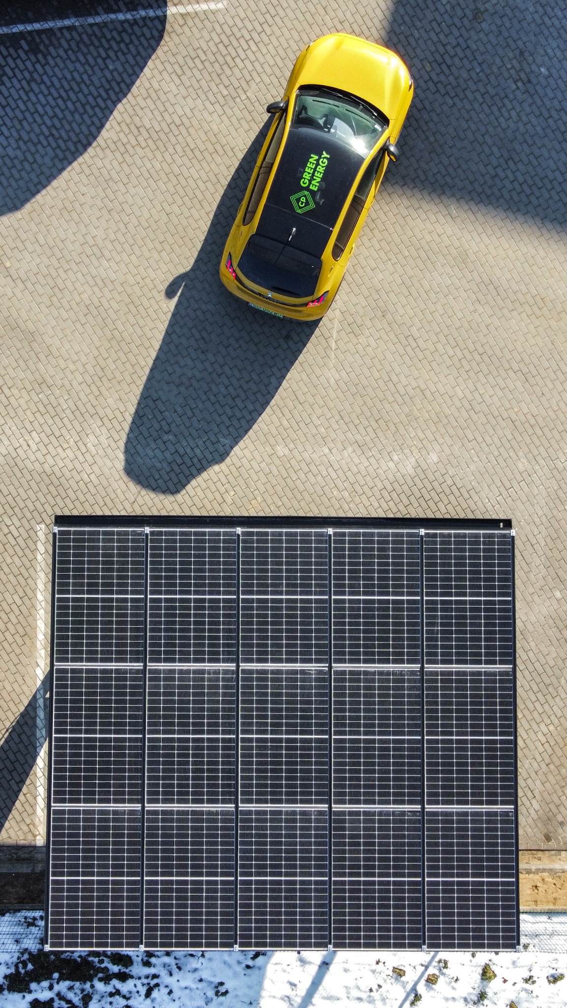 Widok z góry na wiatę fotowoltaiczną (carport) z panelami słonecznymi, obok której stoi żółty samochód z logo 'Green Energy' na dachu, całość na tle kostki brukowej.