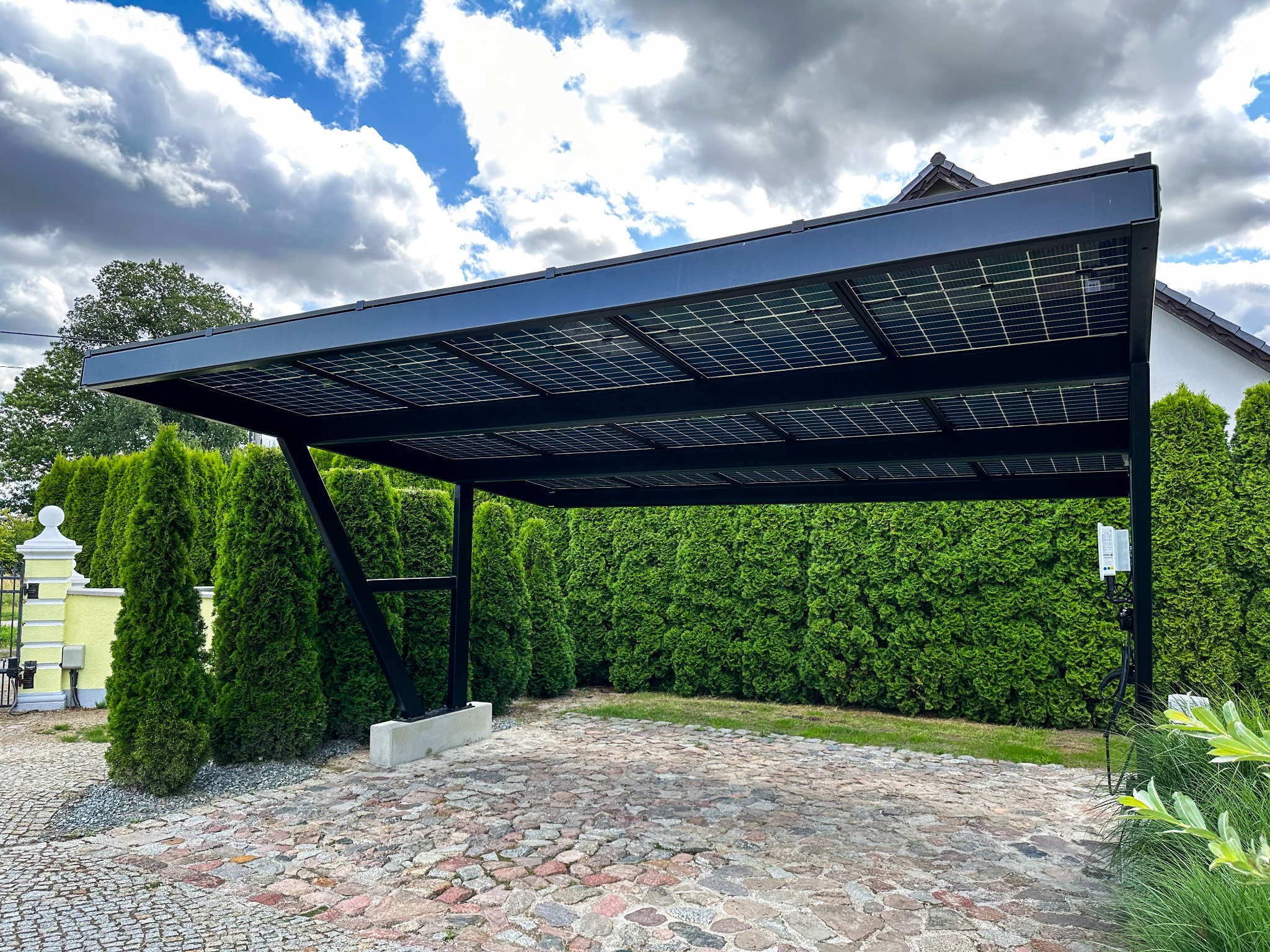 Czarna wiata fotowoltaiczna z panelami słonecznymi, na podjeździe z kostki brukowej, otoczona tujami i elementami ogrodzenia.