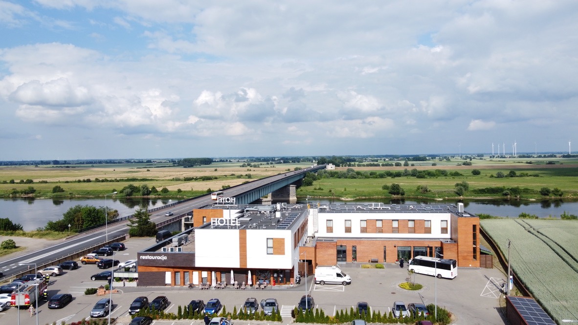 Widok z lotu ptaka na budynek hotelu z panelami słonecznymi na dachu, parkingiem i okolicznym krajobrazem z rzeką, mostem i turbinami wiatrowymi w tle.