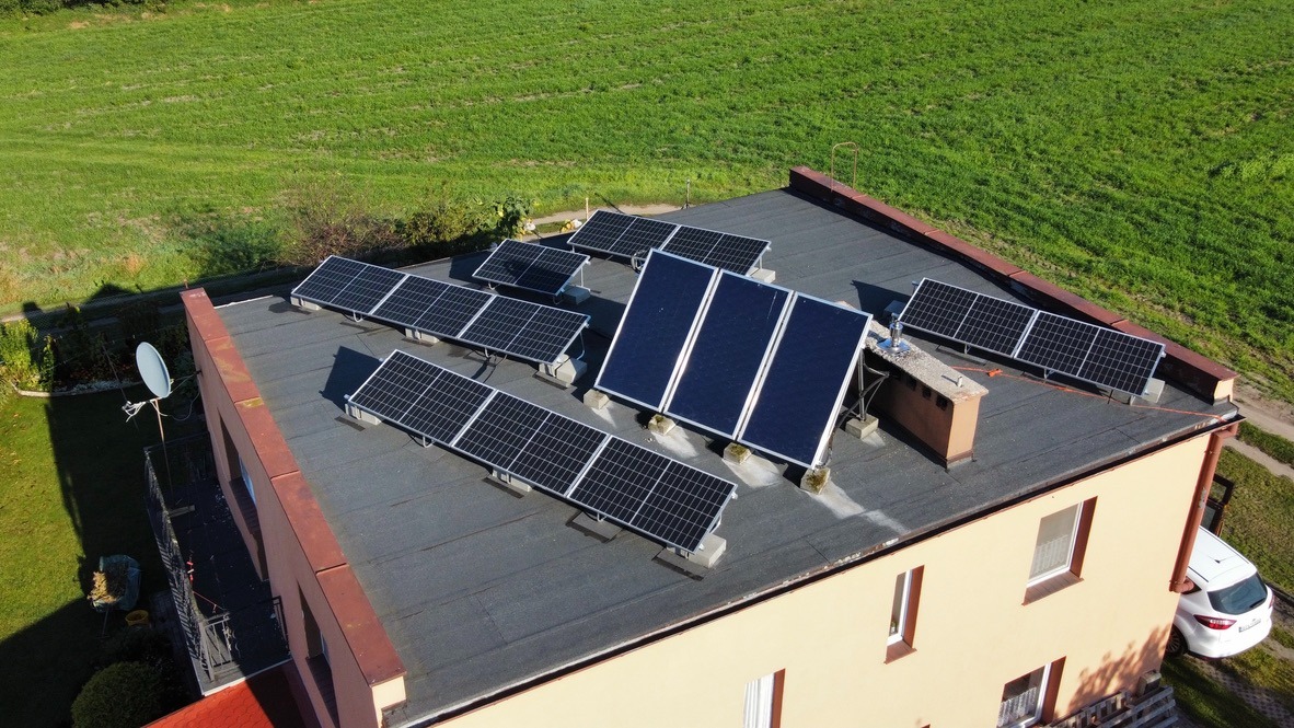 Widok z góry na instalację paneli słonecznych na płaskim dachu domu jednorodzinnego, z widoczną anteną satelitarną i zielonym polem w tle.