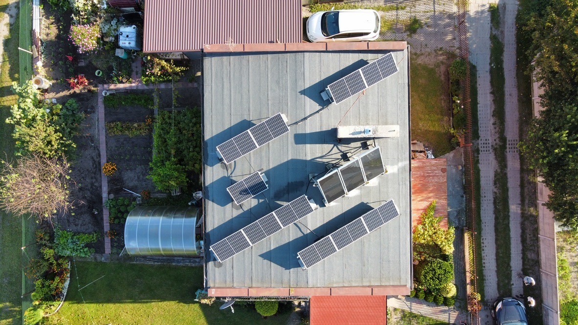 Widok z góry na instalację paneli słonecznych na płaskim dachu budynku jednorodzinnego z ogrodem, obok zaparkowany biały samochód.