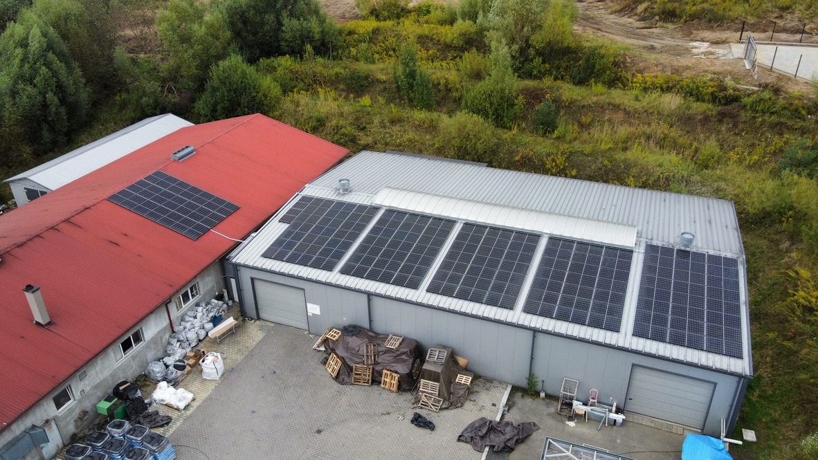 Widok z góry na budynek z dwoma dachami, czerwonym i szarym, oba wyposażone w panele fotowoltaiczne; na szarym dachu cztery rzędy paneli, na czerwonym jeden; na ziemi palety i worki.