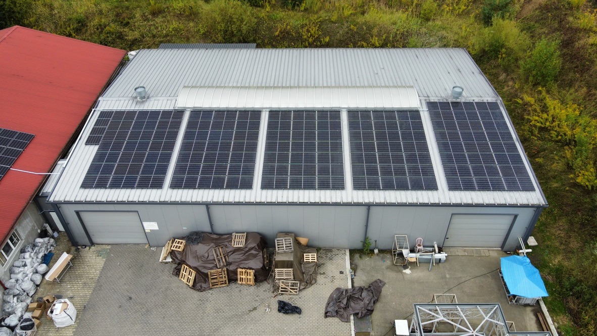 Widok z góry na duży budynek z szarymi ścianami i srebrnym dachem, na którym zamontowano panele słoneczne w kilku sekcjach. Na dachu są też kominy wentylacyjne. Obok znajduje się budynek...