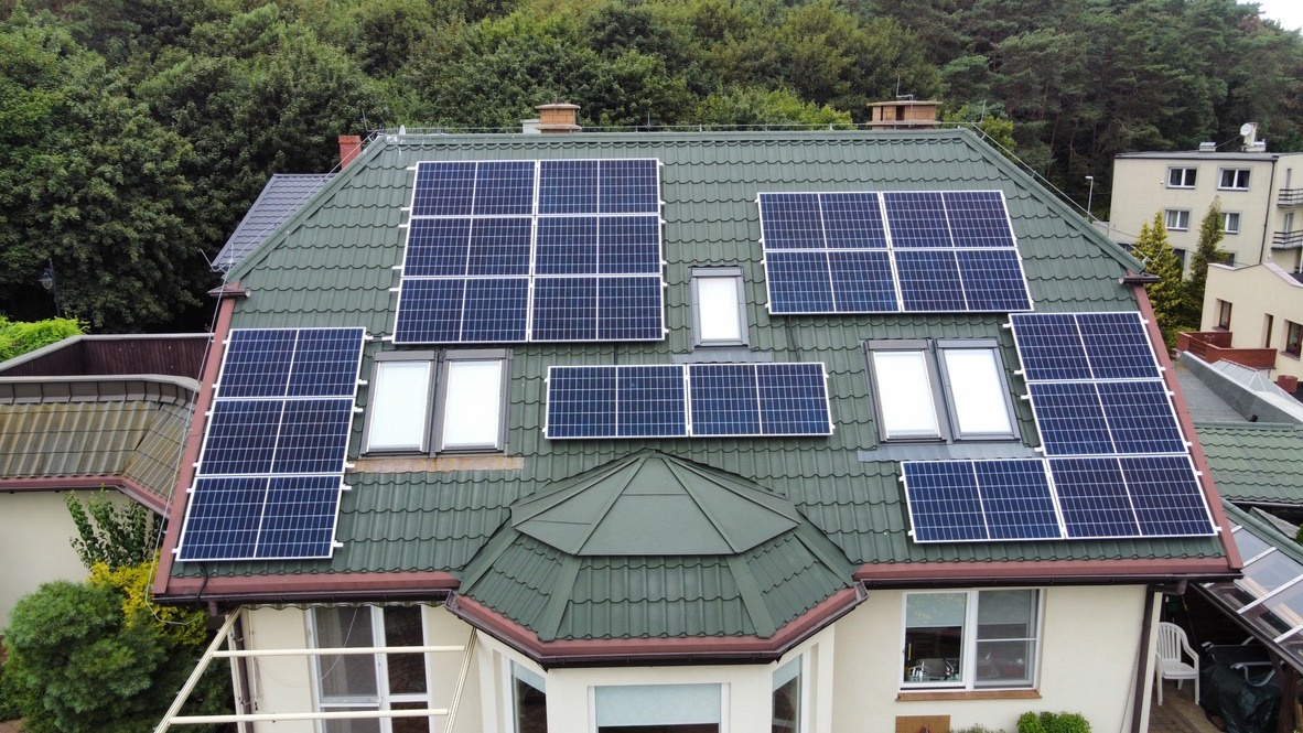 Widok z góry na dach domu z zieloną dachówką, na którym zamontowano panele słoneczne w kilku sekcjach, rozmieszczone wokół okien dachowych; w tle drzewa i budynki.