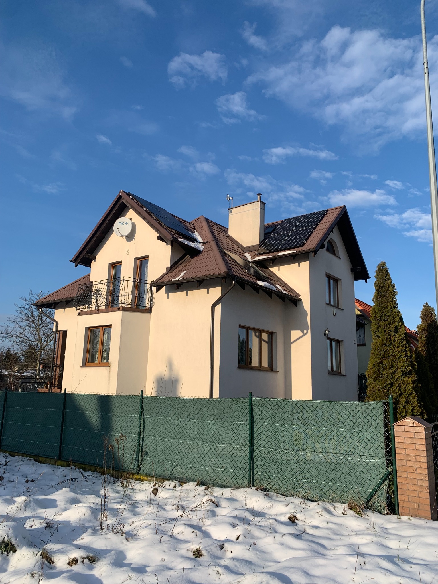 Dwupiętrowy dom z beżową elewacją i brązowym dachem, na którym zamontowane są panele słoneczne. Na dachu widoczna antena satelitarna NC+. Przed domem zielone ogrodzenie z siatki i śnieg.