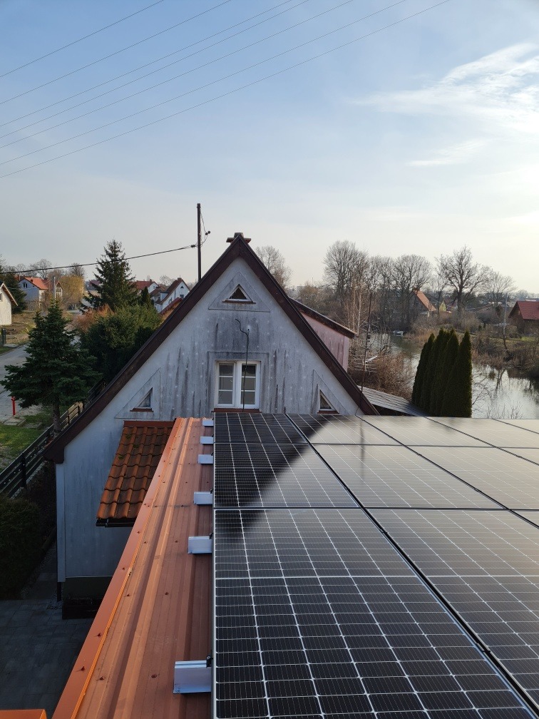 Instalacja paneli słonecznych na dachu dwuspadowym, widok z góry, z widocznymi elementami montażowymi i otoczeniem wiejskim z drzewami i domami.