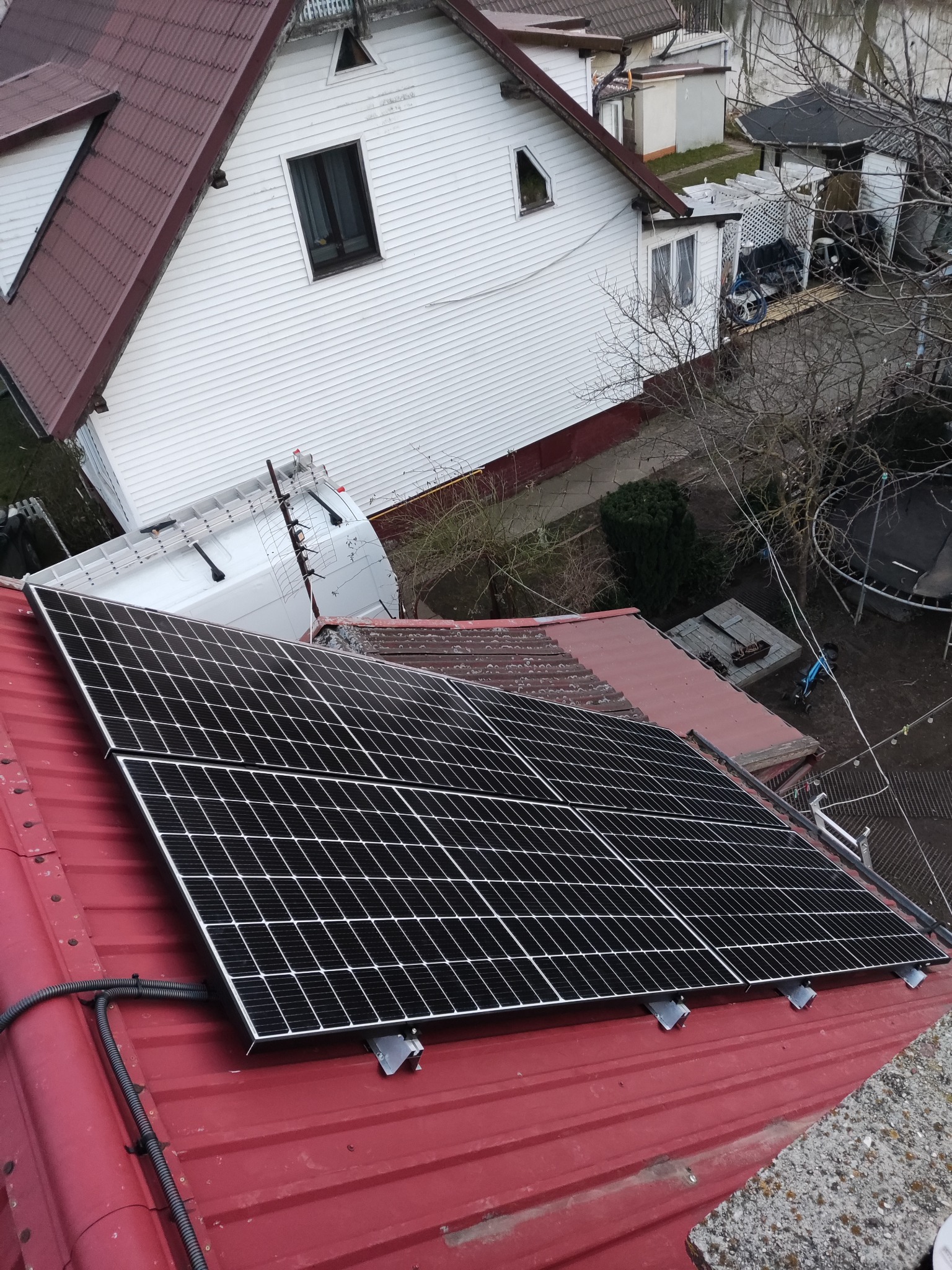 Panele słoneczne zamontowane na czerwonym dachu z blachy falistej, widoczne kable i elementy montażowe, w tle biały budynek i drabina oparta o dach.