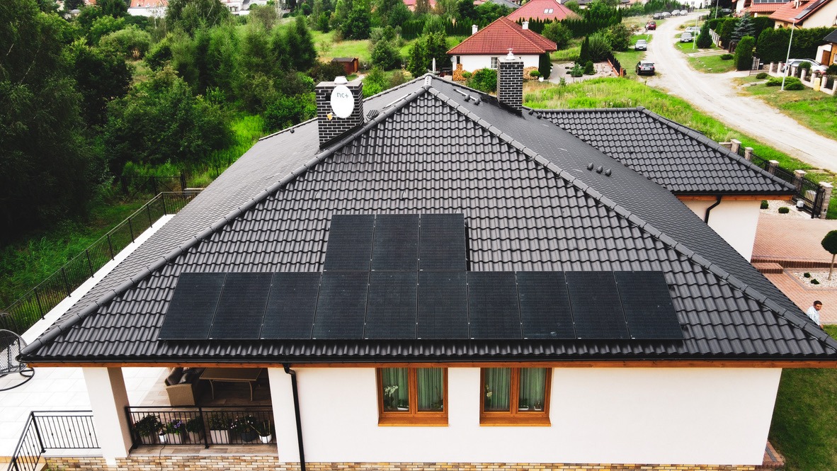 Widok z góry na dom z ciemną dachówką i panelami słonecznymi. Panele ułożone w dwóch rzędach, z satelitą na kominie w tle.