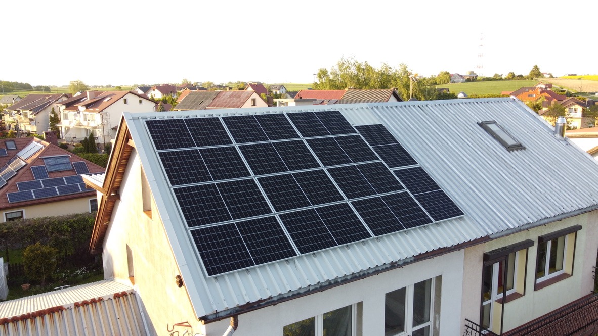 Instalacja fotowoltaiczna z panelami ułożonymi w prostokątny wzór na falistym, metalowym dachu domu jednorodzinnego z oknem dachowym, widok z lotu ptaka na tle zabudowy wiejskiej.