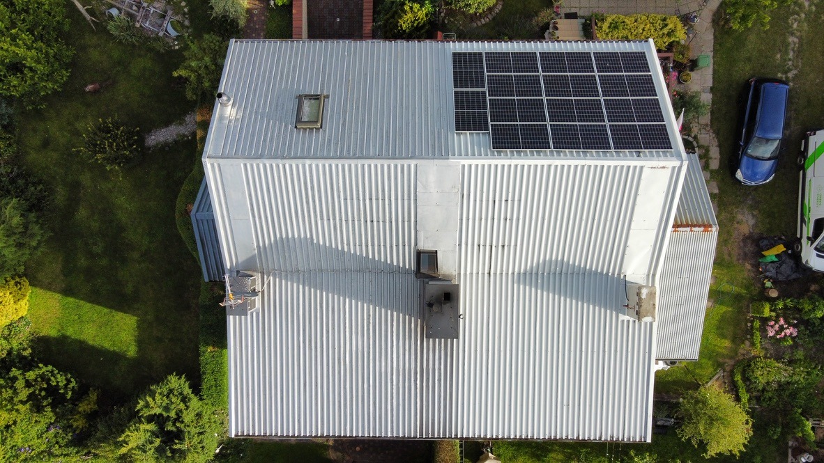 Widok z góry na dom z blachy falistej z zamontowanymi panelami słonecznymi po prawej stronie dachu, obok okna dachowego, na zadbanym trawniku z krzewami i drzewami, obok zaparkowany niebieski...