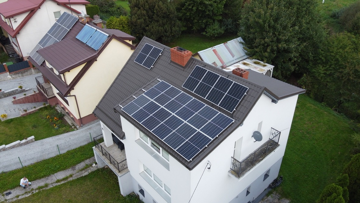 Widok z góry na dom jednorodzinny z brązowym dachem, na którym zamontowane są panele fotowoltaiczne w dwóch sekcjach; na sąsiednich budynkach również widać instalacje solarne, a na trawniku siedzi...
