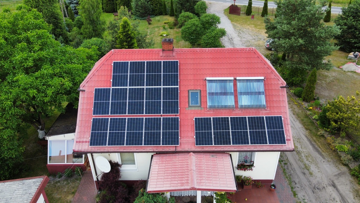 Widok z lotu ptaka na dom z czerwonym dachem, na którym zamontowano panele słoneczne i kolektory słoneczne. Dodatkowo widoczny komin z cegły i okno dachowe.