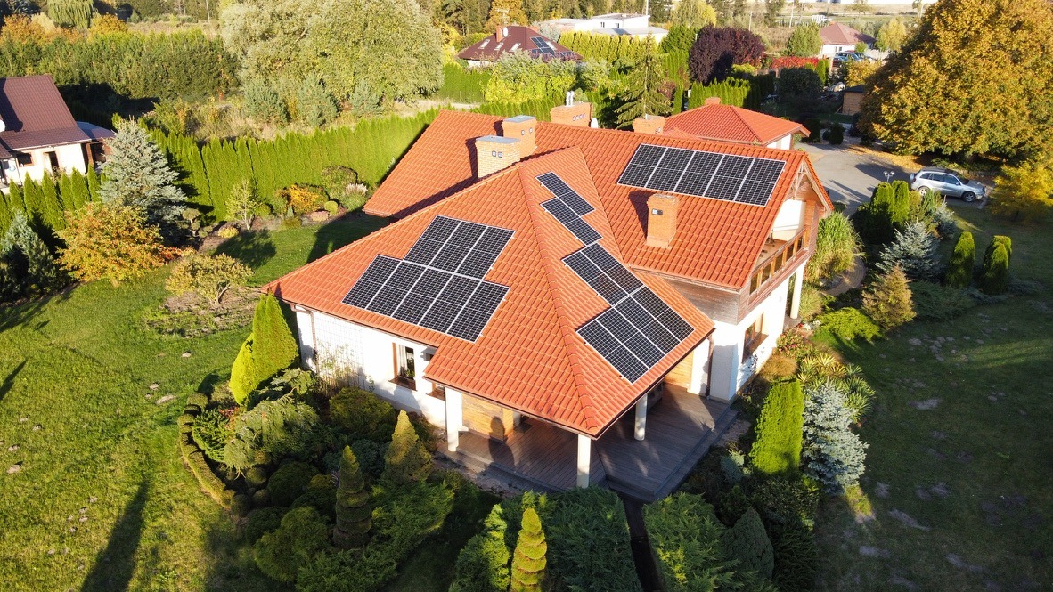 Widok z lotu ptaka na dom jednorodzinny z pomarańczową dachówką, na której zamontowane są panele słoneczne. Wokół domu bujna zieleń, w tle widoczne inne domy i drzewa.