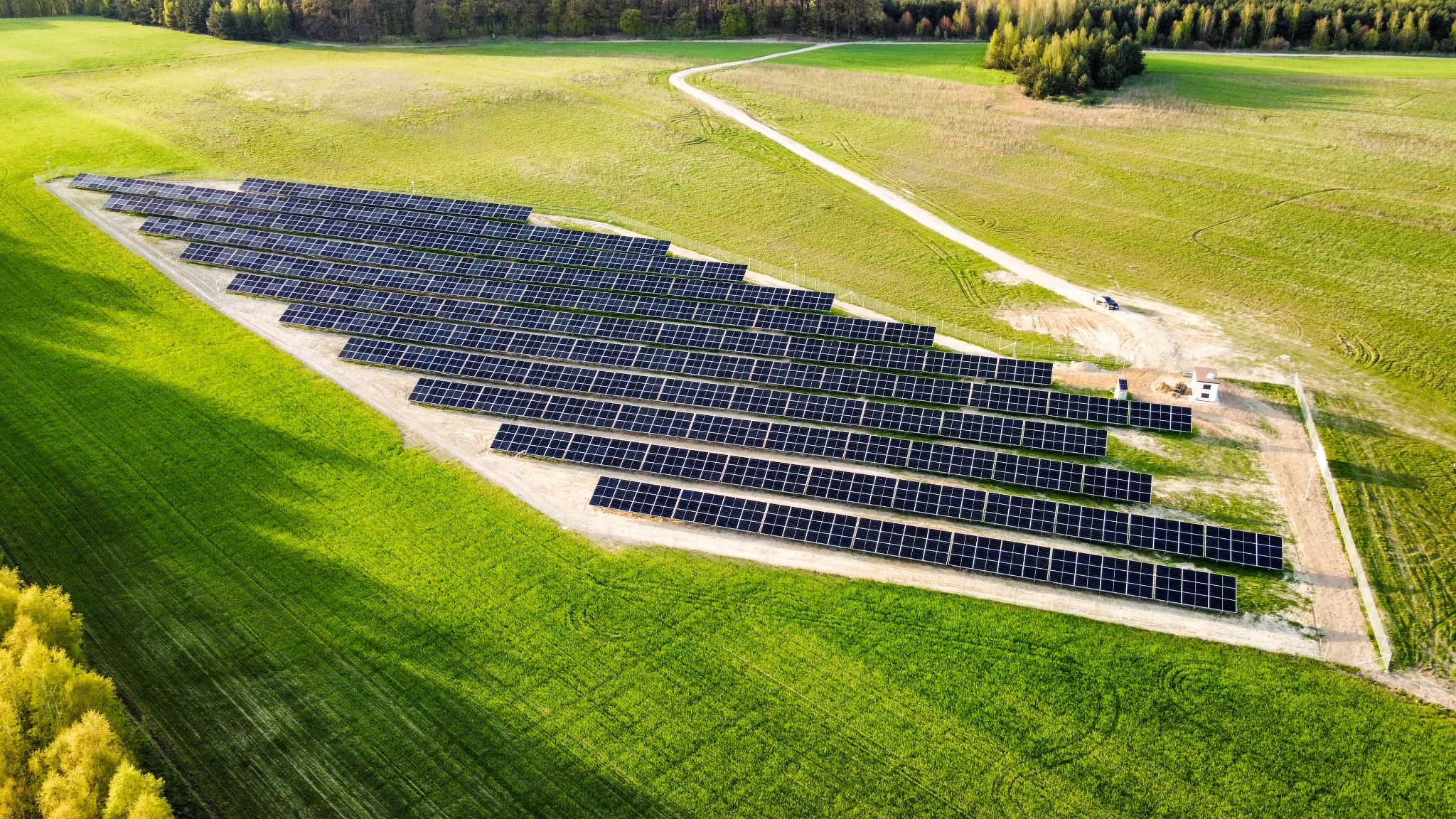 Widok z lotu ptaka na rozległą instalację paneli słonecznych na gruncie, otoczoną zielonym polem i drzewami, z widoczną drogą dojazdową i małym budynkiem technicznym.