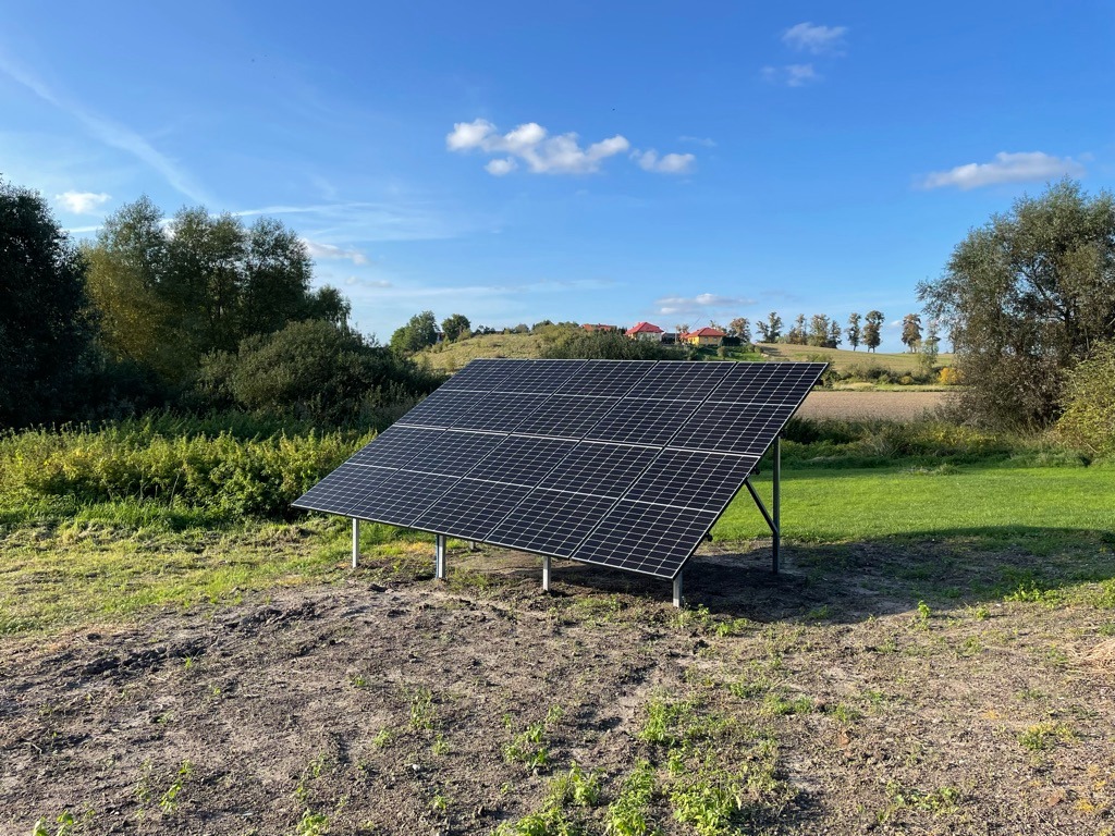 Instalacja fotowoltaiczna na gruncie z widocznymi panelami, metalową konstrukcją wsporczą i otaczającym krajobrazem polnym z drzewami w tle pod błękitnym niebem z chmurami.